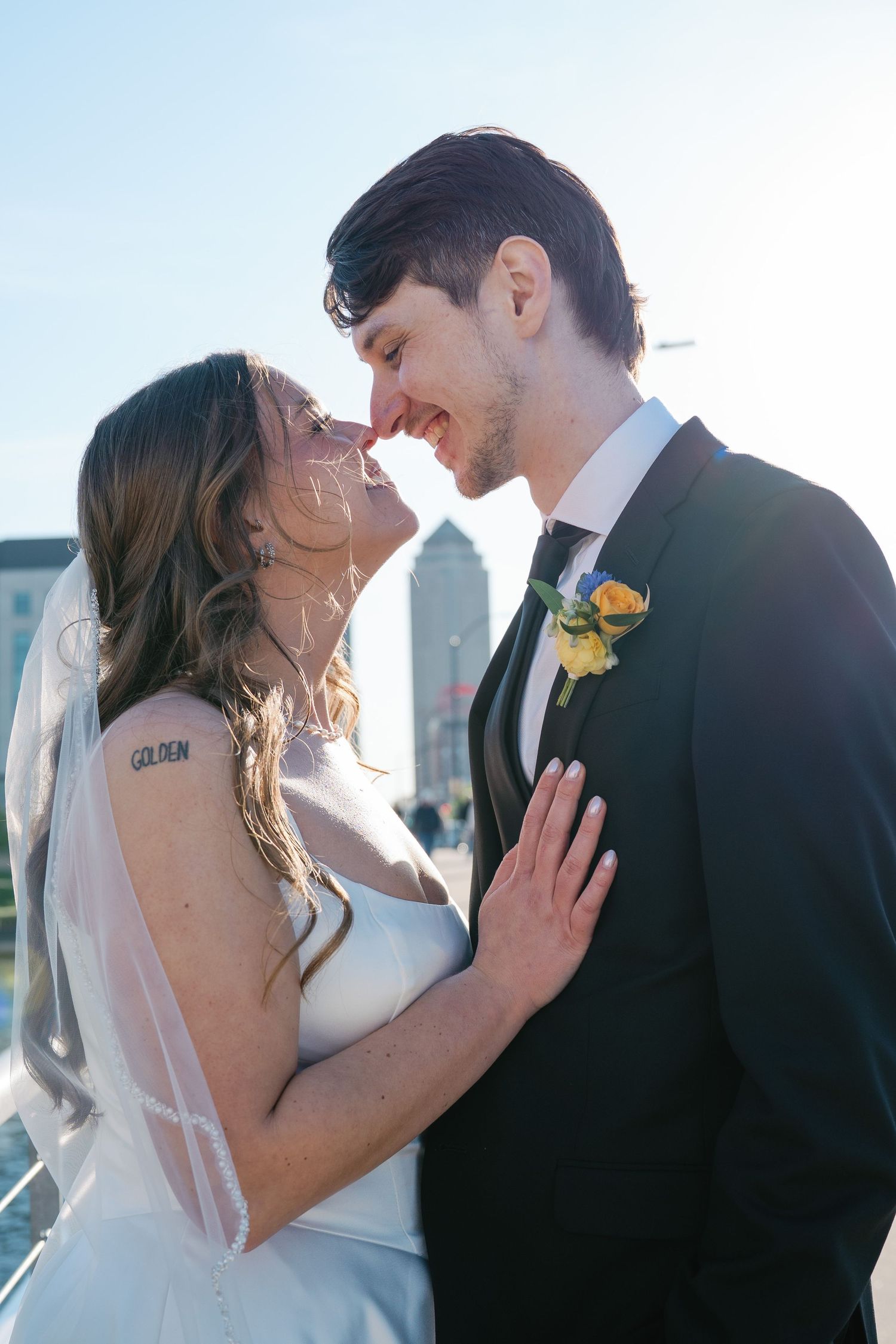 Bride and groom share a romantic moment, nearly kissing outdoors in golden sunlight, with city skyline in background.