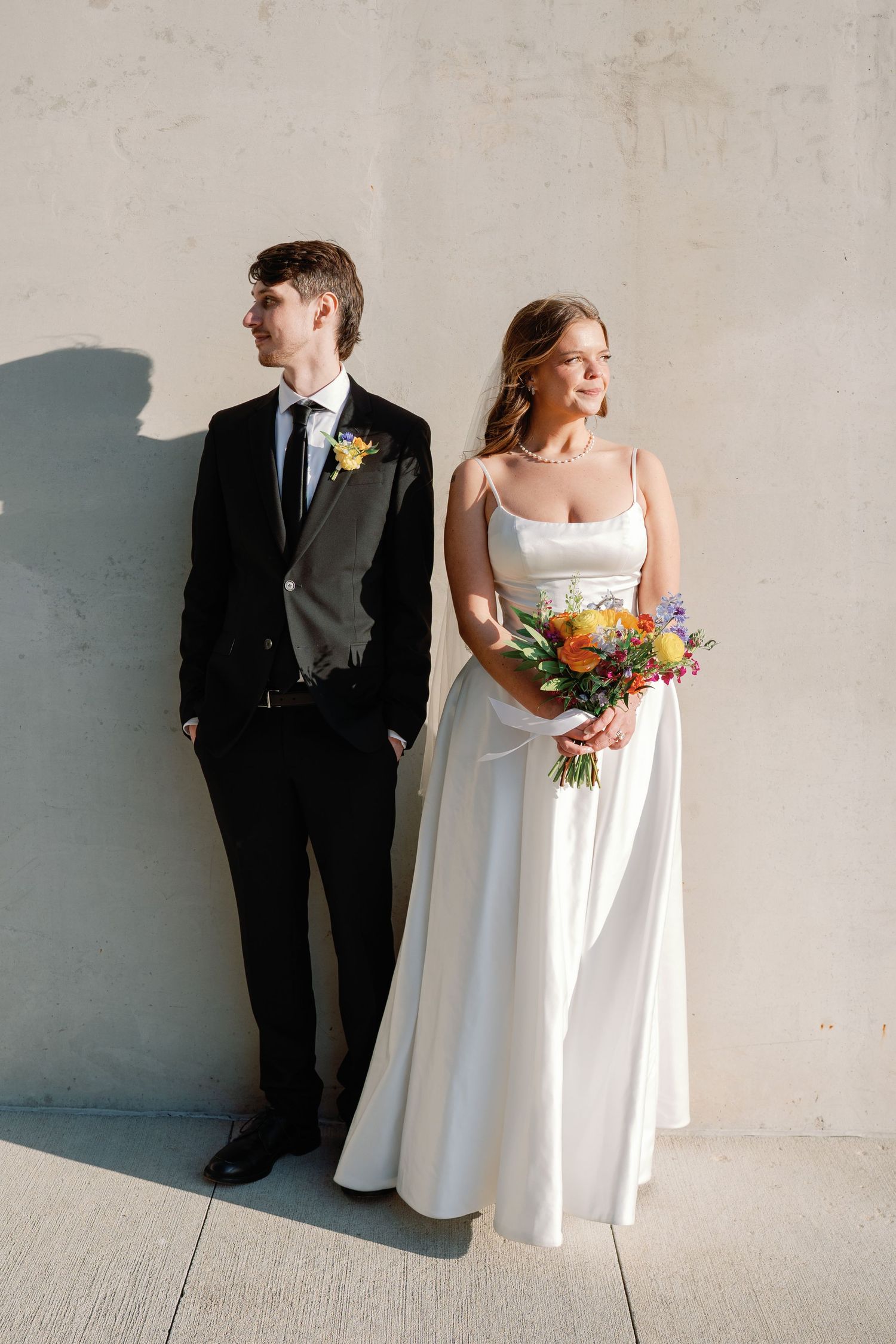 Bride in white gown holding colorful bouquet stands with groom in black suit against concrete wall.