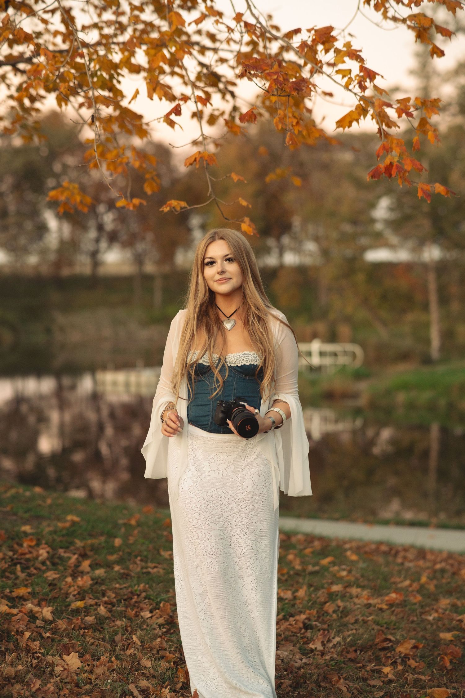 Woman in white sweater poses with camera among autumn leaves.