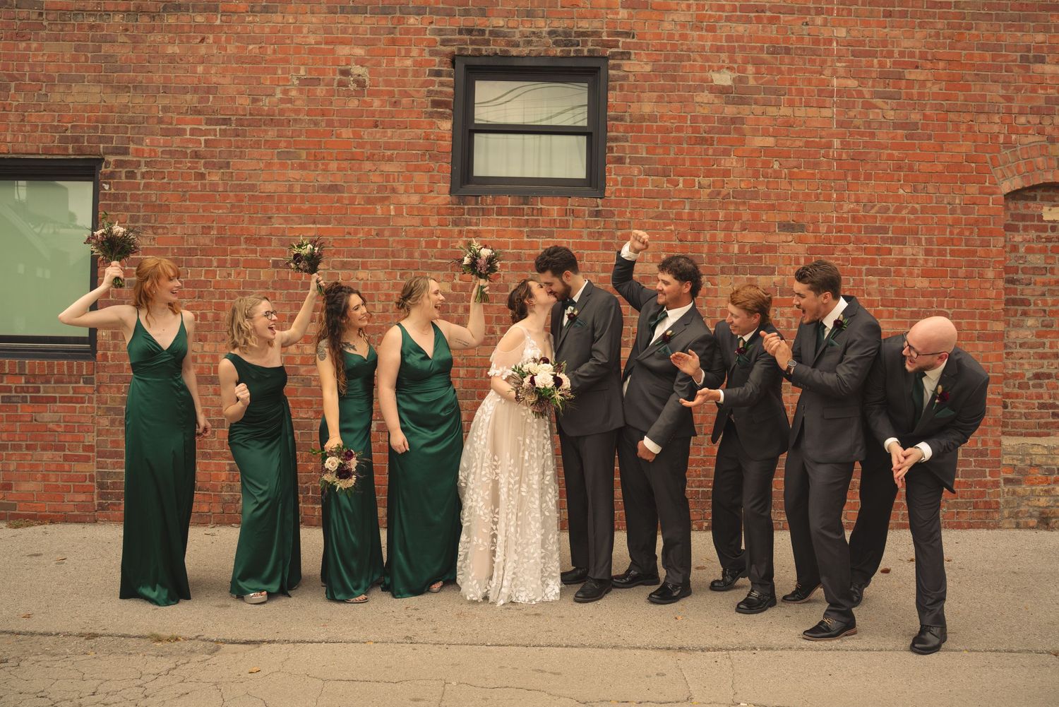 Wedding party poses against rustic brick wall wearing emerald green dresses and dark suits.
