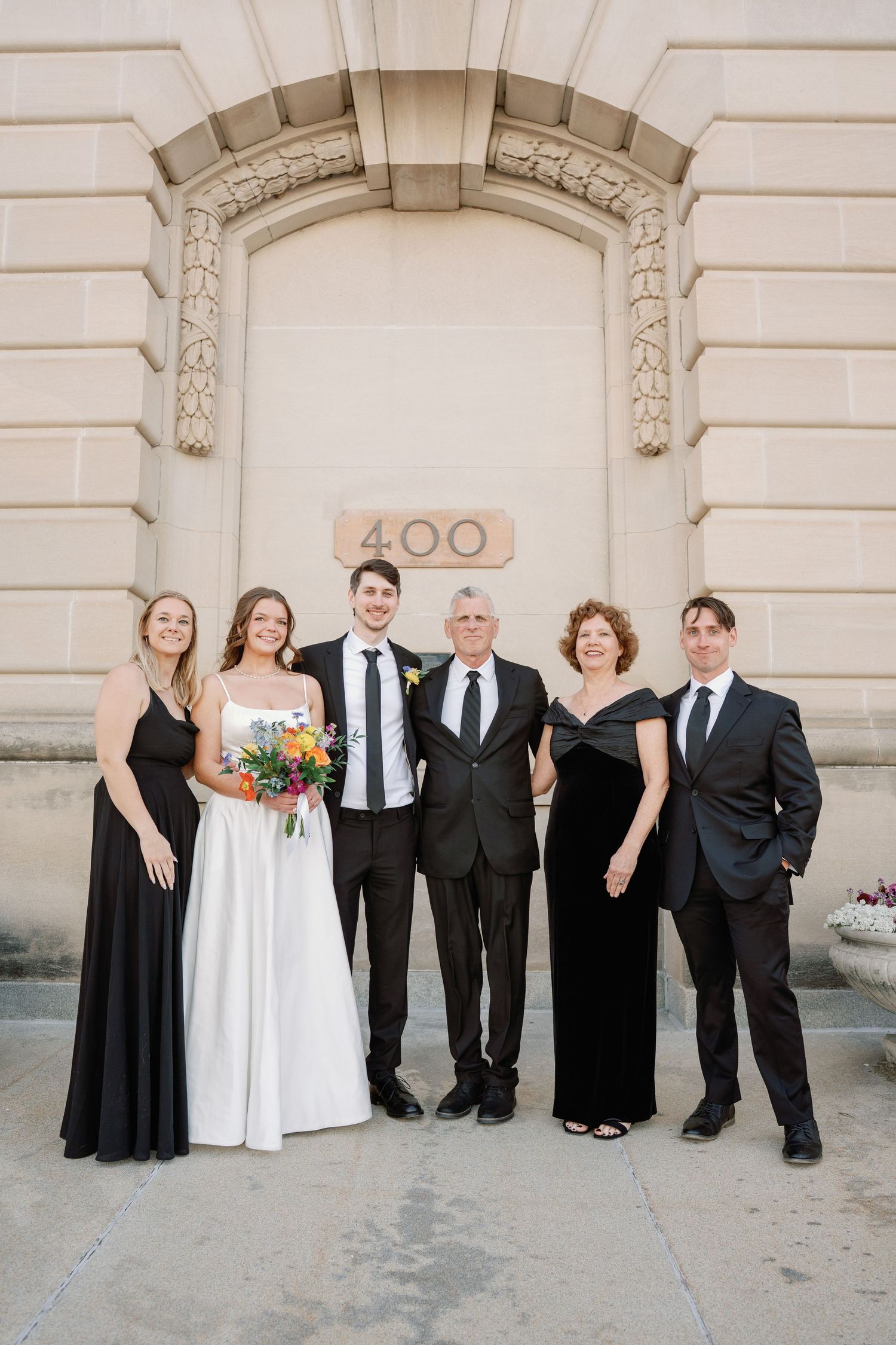 Wedding party of six poses outside building 400, bride in white gown holding colorful bouquet with groom and family.