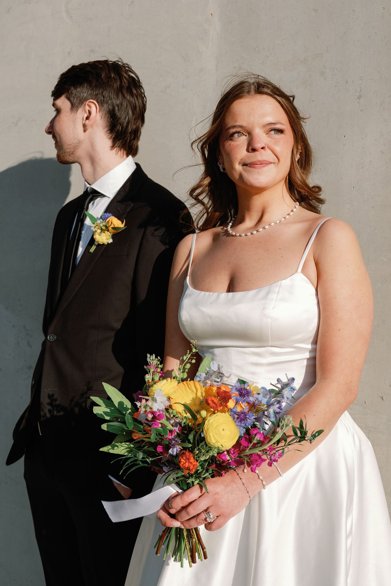 Bride in white dress holds colorful bouquet beside groom in black suit against gray wall.