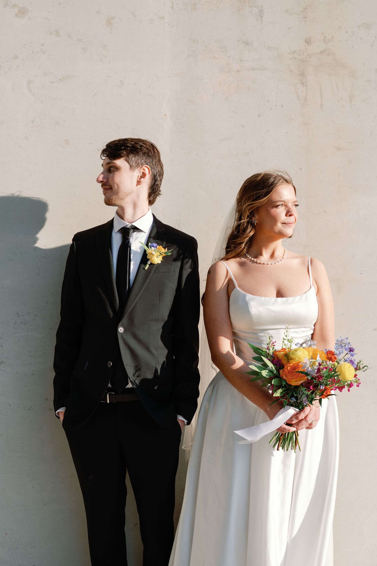 Bride in white gown holding colorful bouquet stands beside groom in black suit with yellow boutonniere against gray wall.