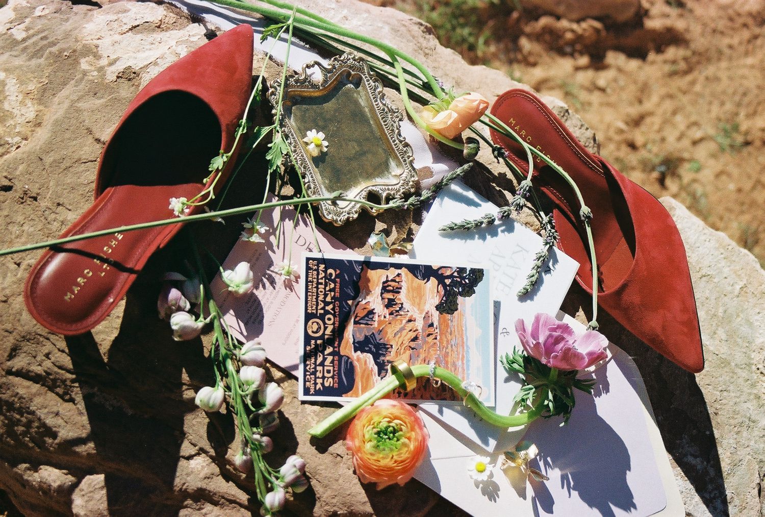 Red suede heels, floral garlands, vintage mirror, and a magazine arranged on rocky surface.
