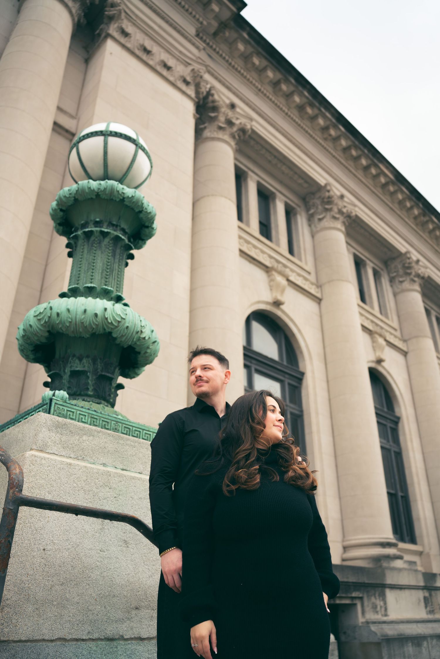 Couple in black outfits posing near ornate green lamp post outside a classic beige stone building with columns.