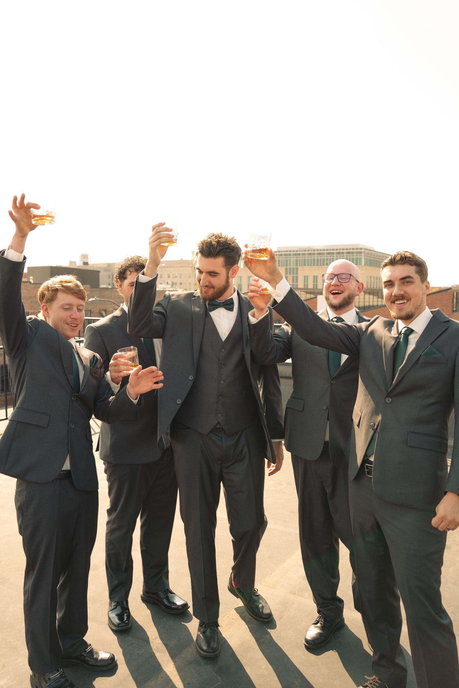 Groomsmen in black suits posing playfully in a line outdoors.