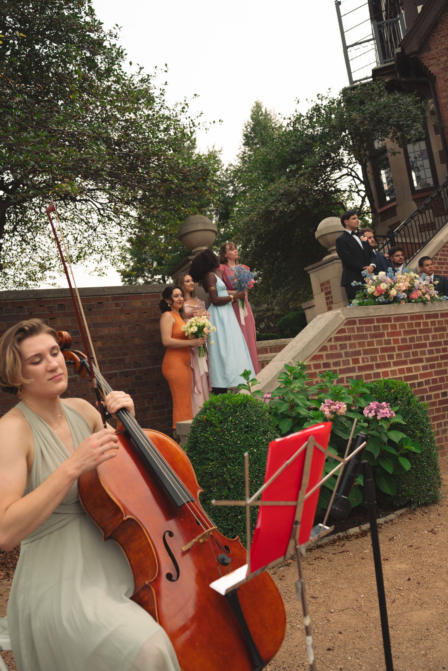 A cellist performs outdoors against a brick wall backdrop during a garden wedding ceremony.