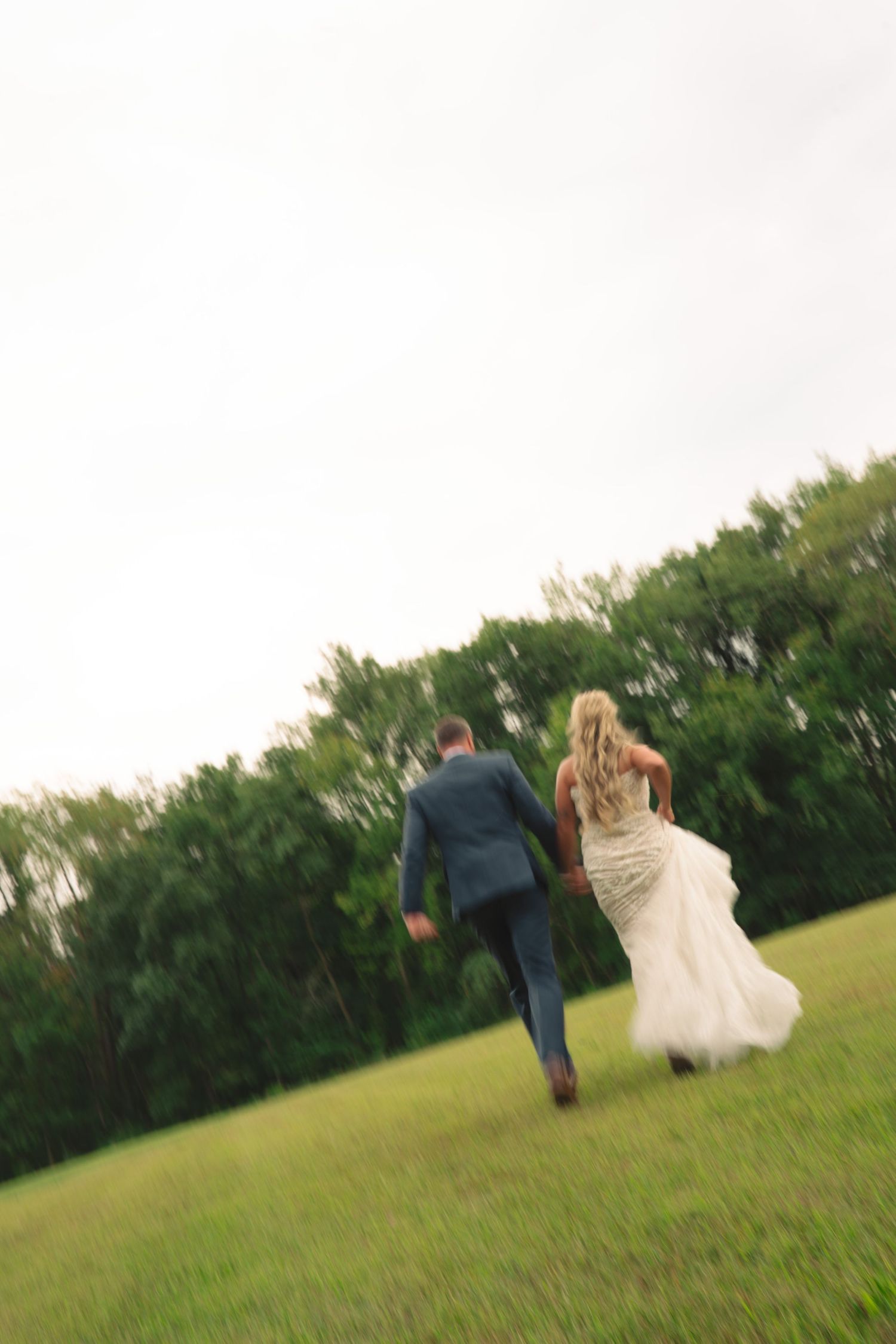 Wedding couple running playfully through a green field near trees.