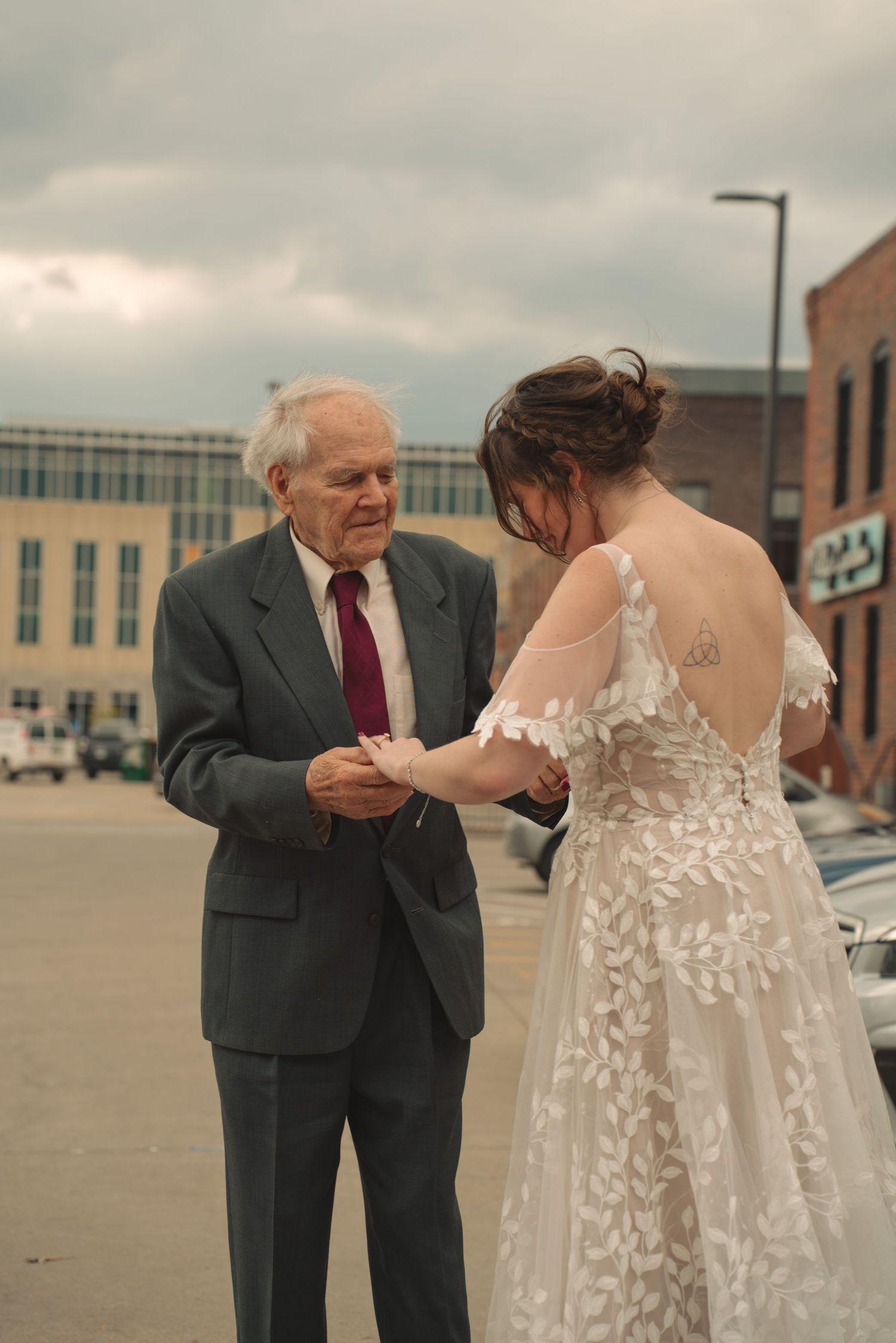 Bride in lace gown shares tender moment with elderly guest in grey suit outdoors.