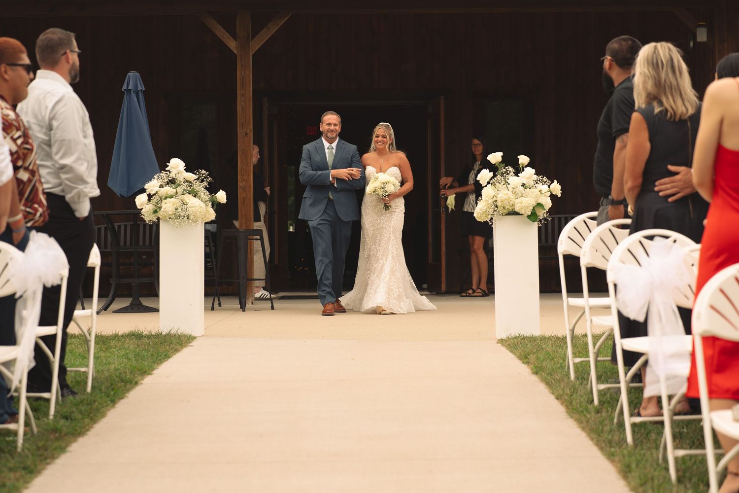 White folding chairs line an outdoor wedding ceremony aisle under a covered pavilion.
