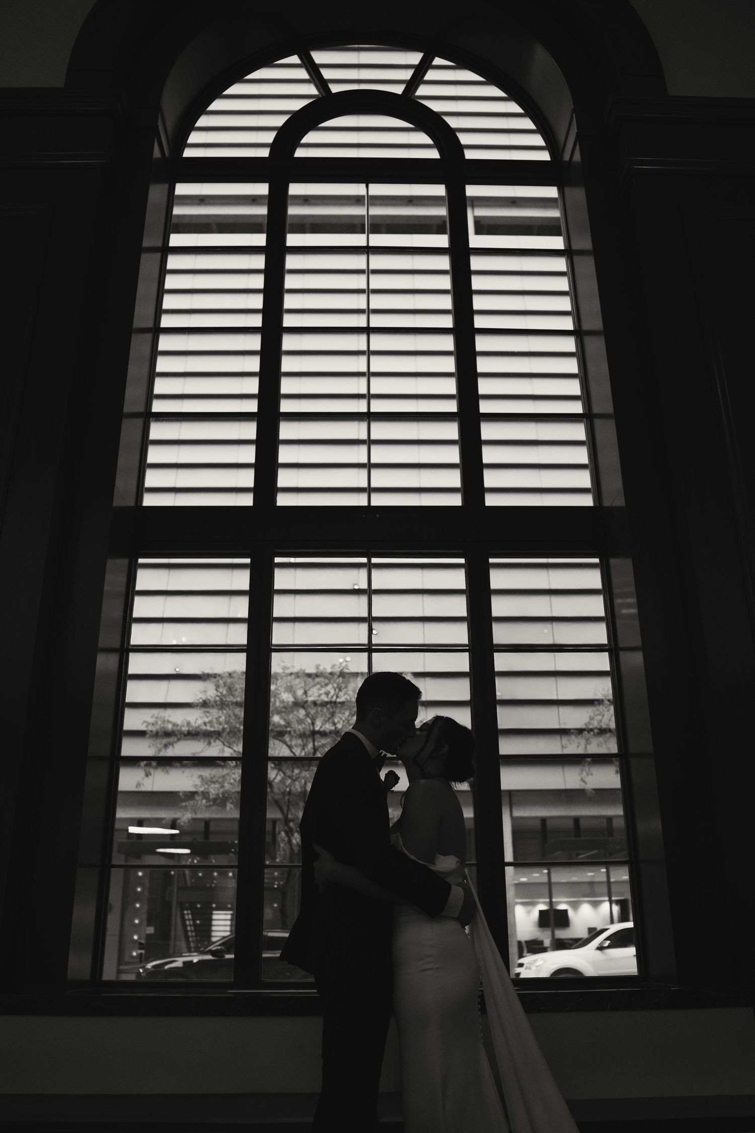 Silhouetted couple in romantic embrace against bright arched window with horizontal blinds creating dramatic lighting.
