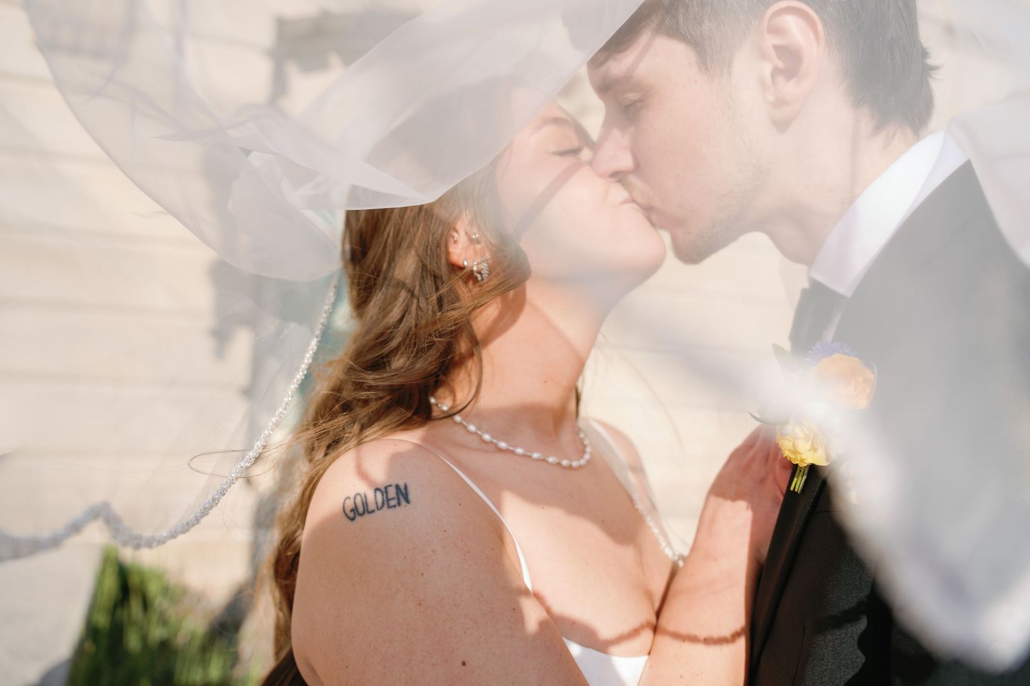 Bride and groom share a romantic kiss under a flowing wedding veil, bride wearing pearl necklace and Golden tattoo.