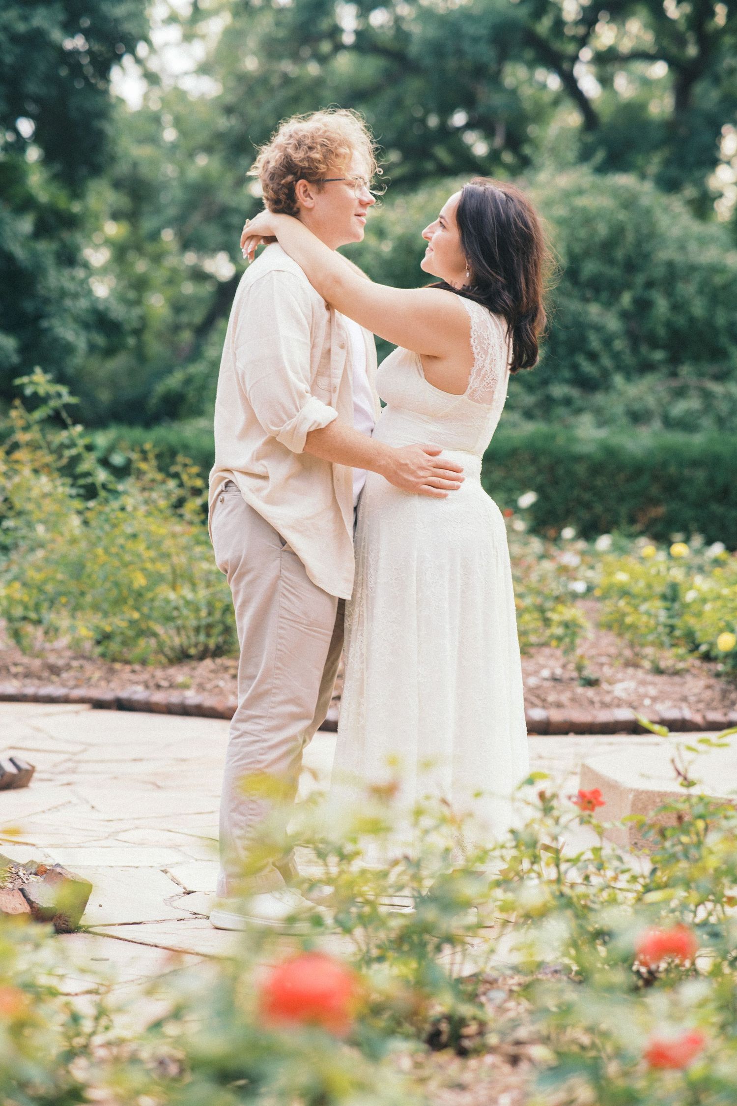 Couple embracing and smiling at each other in a lush garden, woman in white dress, man in casual beige outfit.