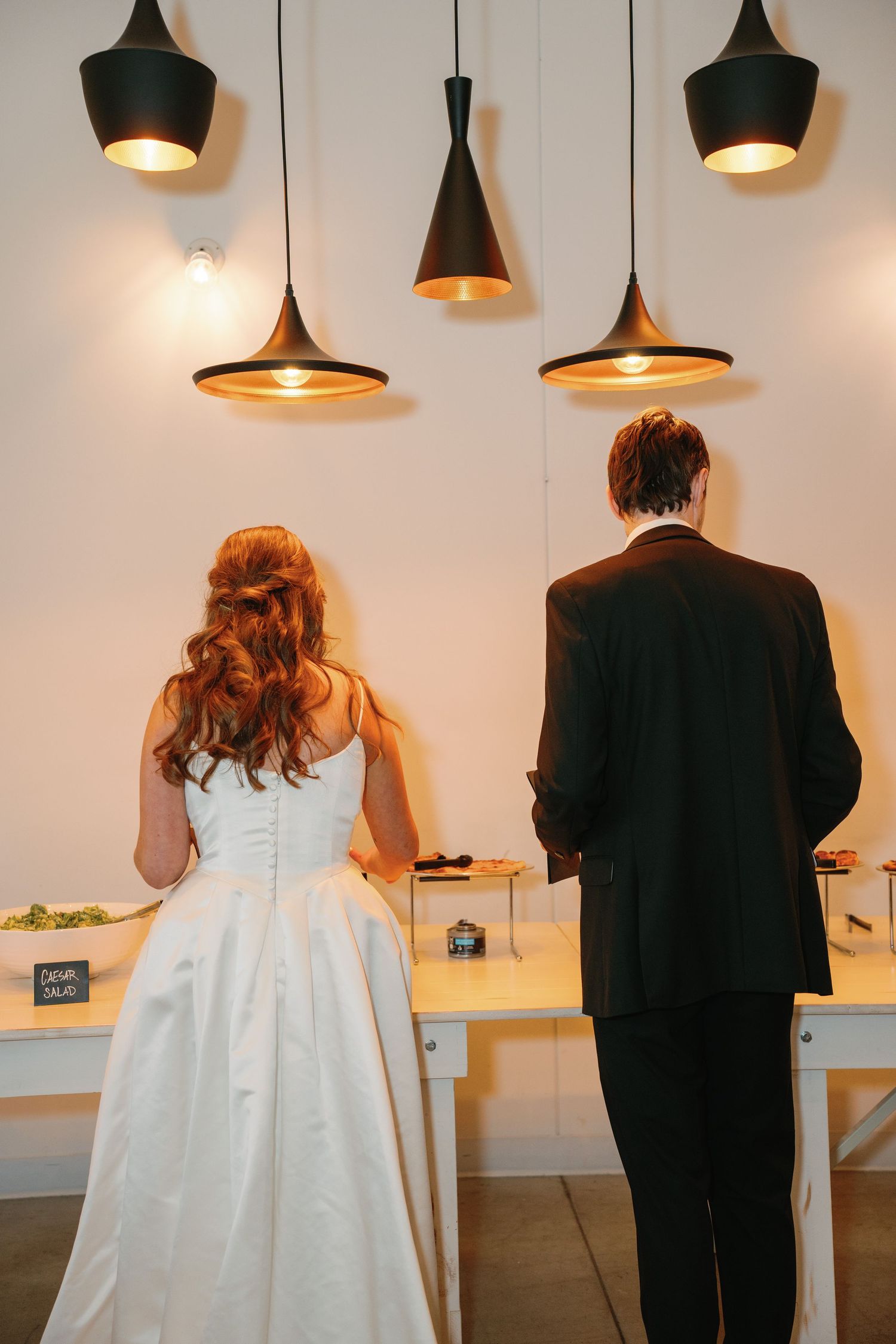 Bride and groom stand at a wedding reception buffet table beneath modern black pendant lights.