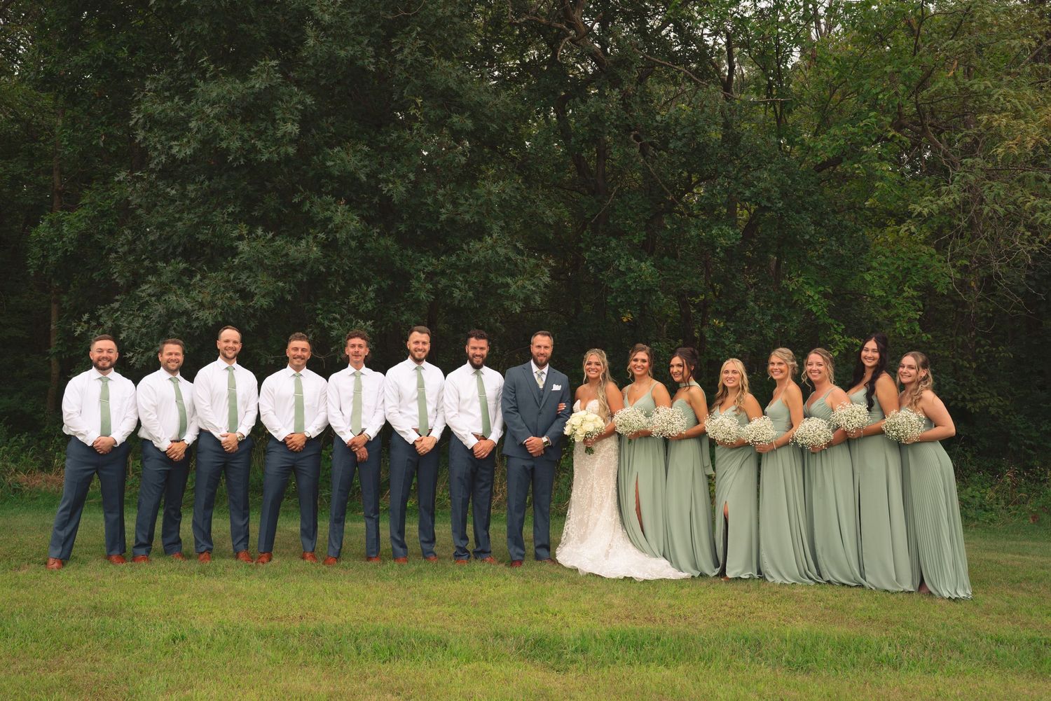 Full wedding party lineup with bridesmaids in sage green and groomsmen in navy against natural backdrop.