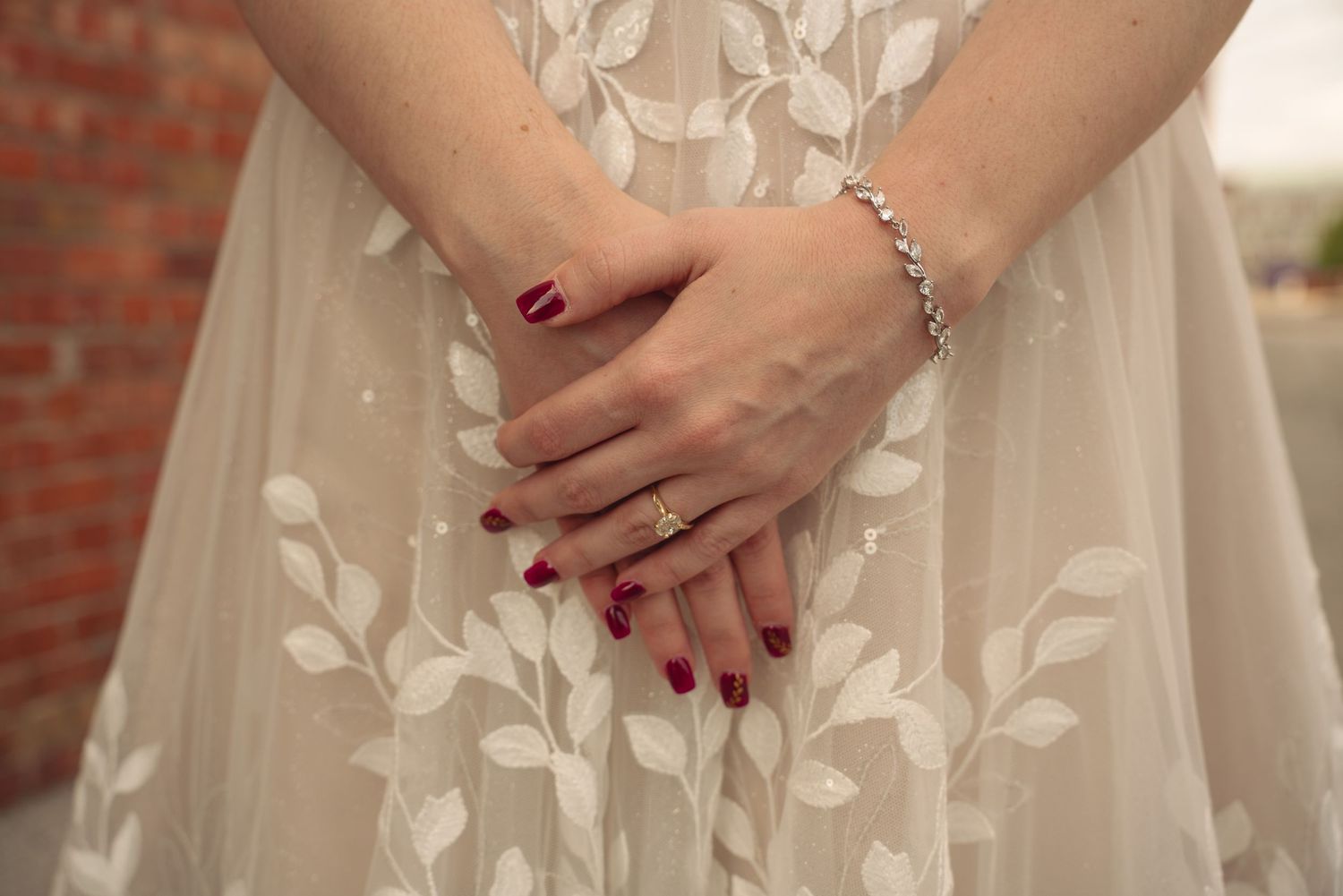 Close-up of bride's hands with red manicure resting on white floral lace wedding dress.