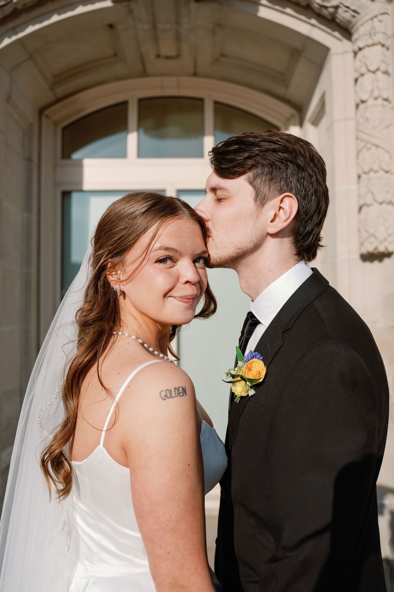 Groom in black suit kissing bride on cheek as she smiles, standing before elegant stone building on wedding day.