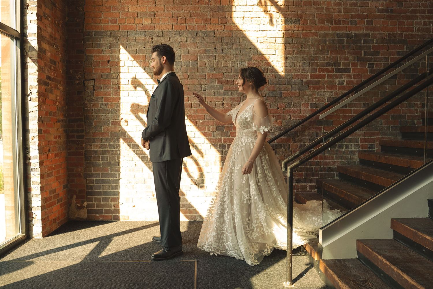 Bride and groom stand in a sunlit industrial venue with exposed brick walls and wooden stairs.