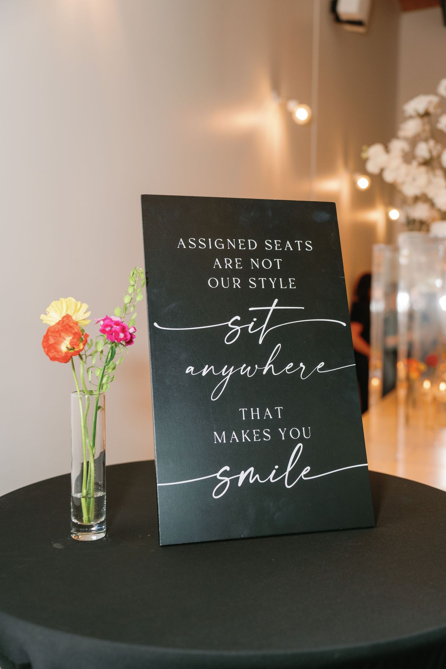 Black wedding sign reading sit anywhere that makes you smile, displayed beside colorful flowers in a glass vase.