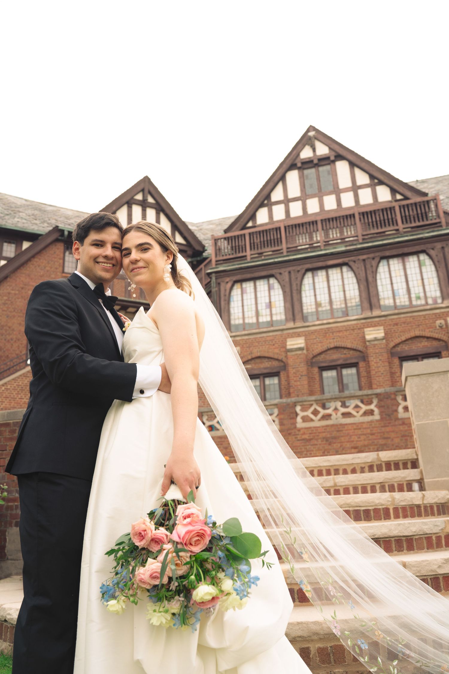 Wedding couple standing on steps of historic Tudor-style building with flowing veil.