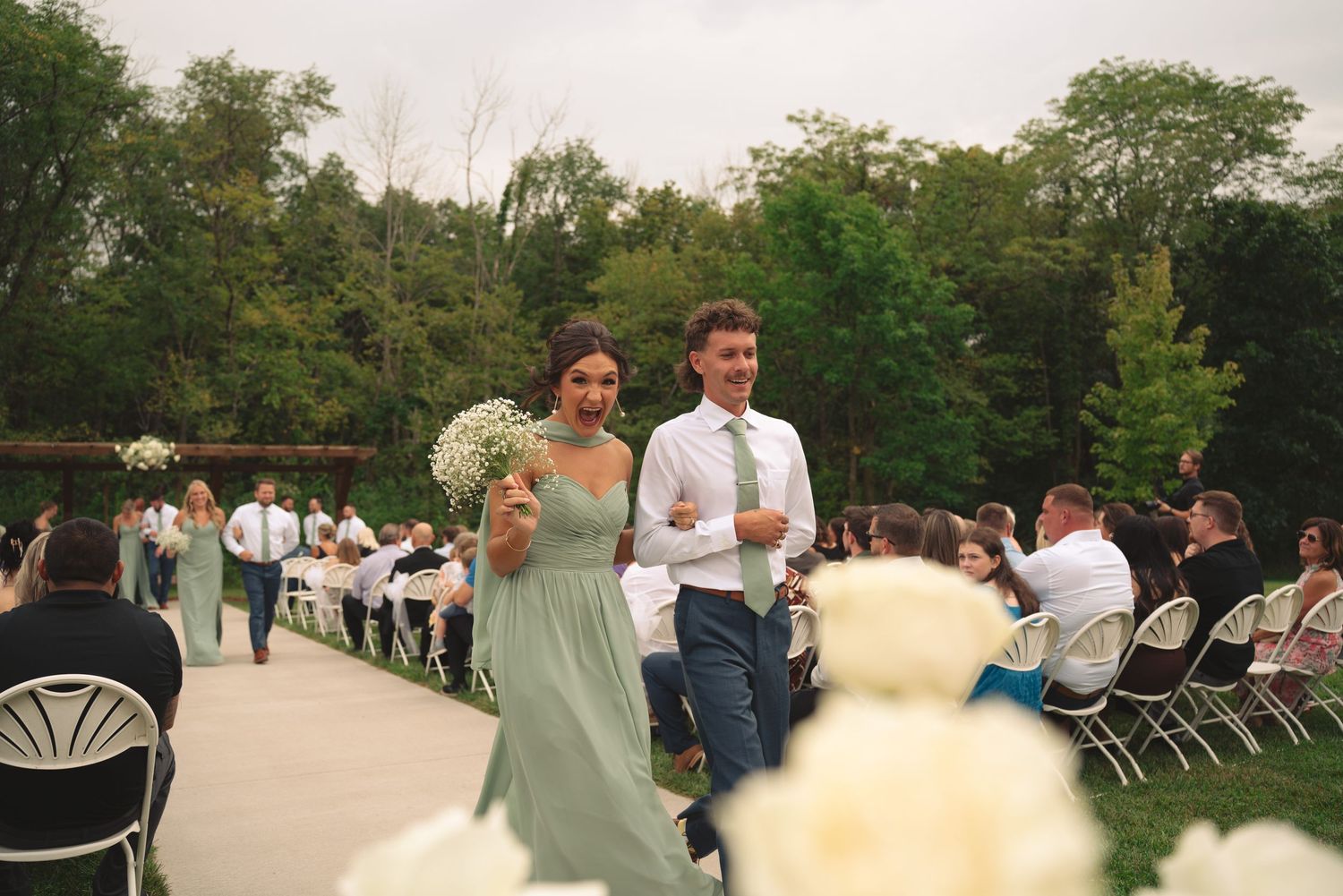 Wedding ceremony in progress with guests seated on white chairs in garden setting.