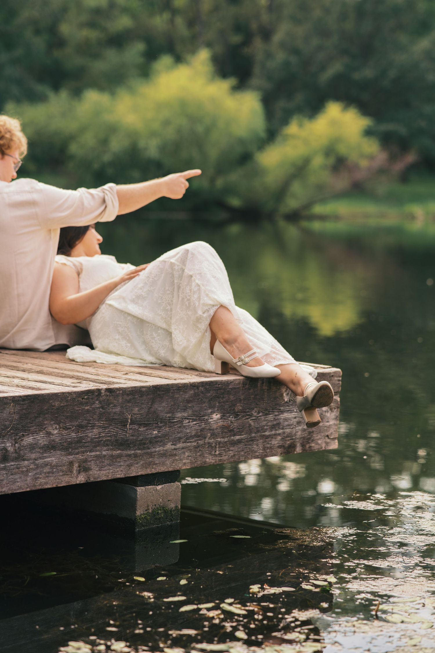 Couple sitting on a wooden dock by a serene lake, surrounded by lush green trees, pointing into the distance.