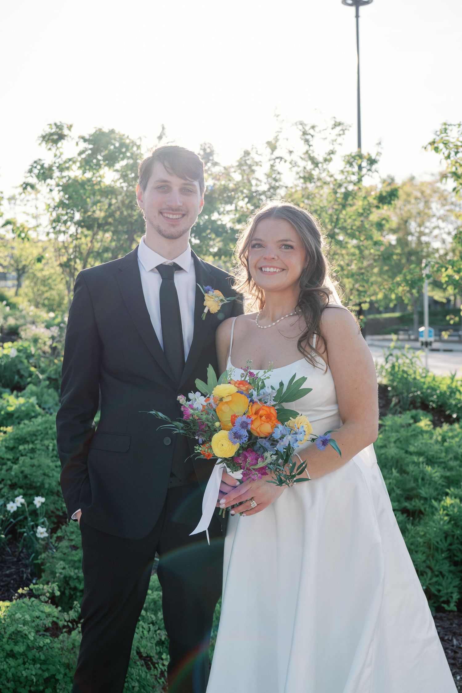 Bride and groom pose outdoors in wedding attire, she holds a colorful bouquet, surrounded by lush greenery.