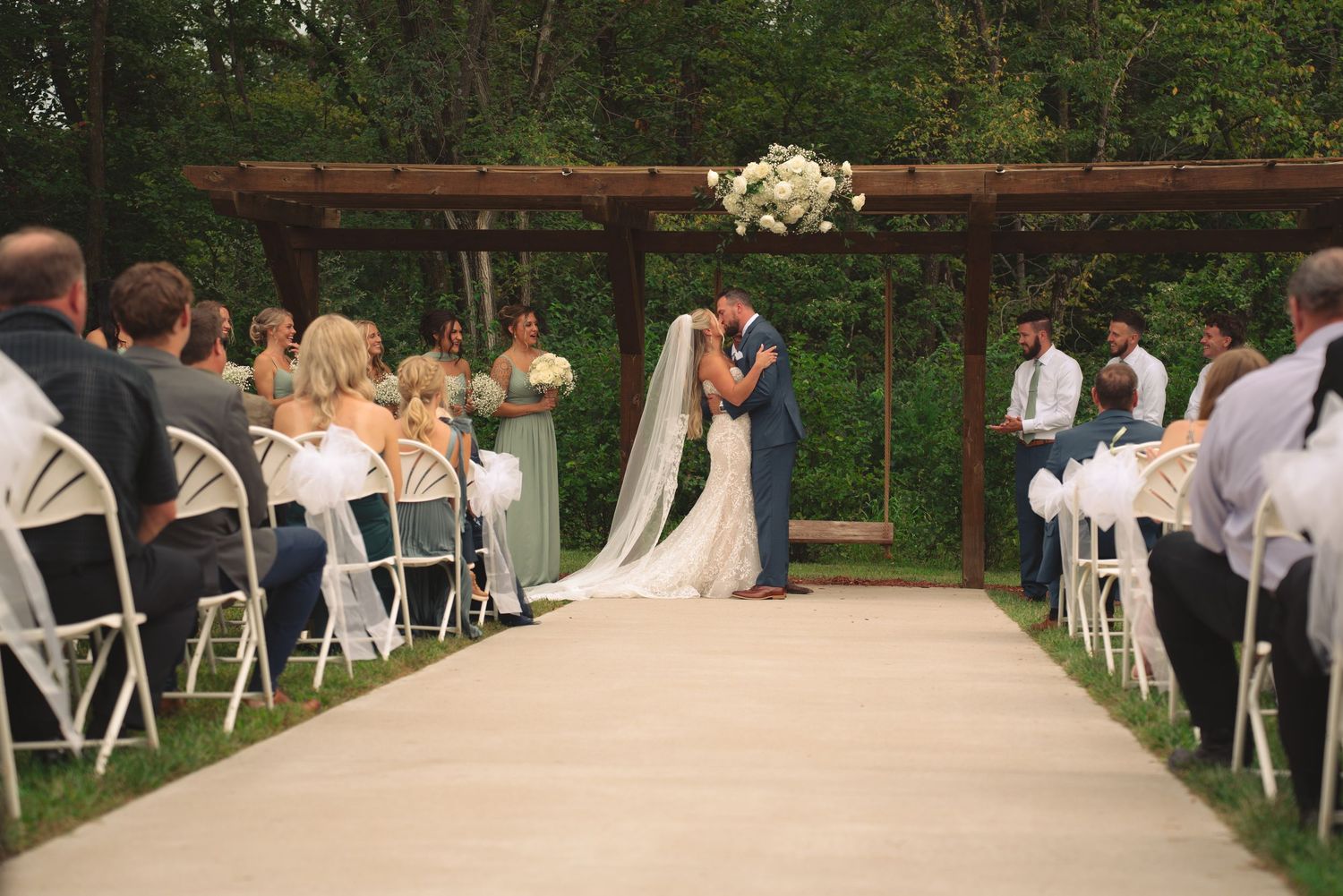 Outdoor wedding venue with wooden pergola and rows of white chairs surrounded by trees.