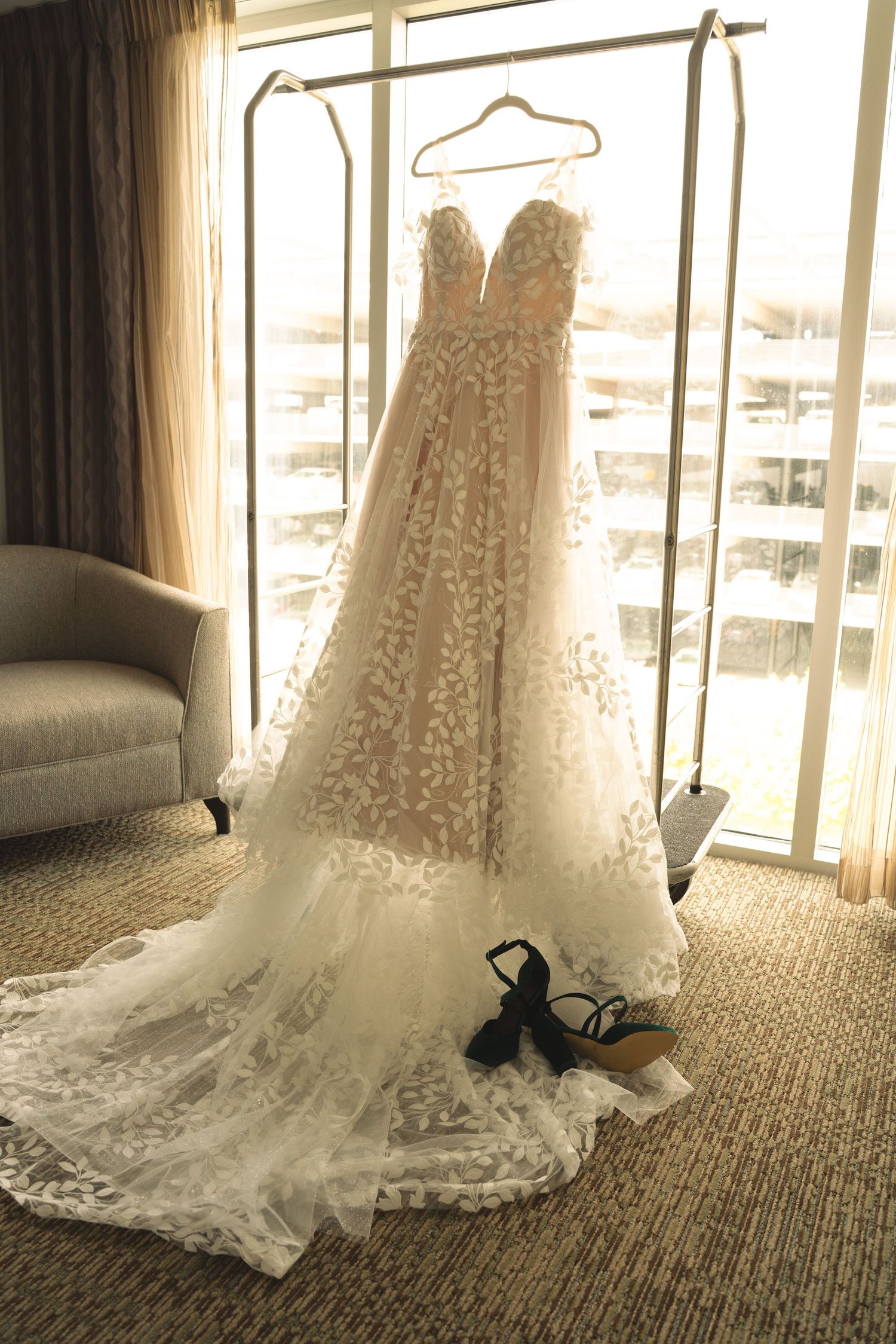 An elegant bridal gown with intricate lace details spread out on hotel room carpet near window.