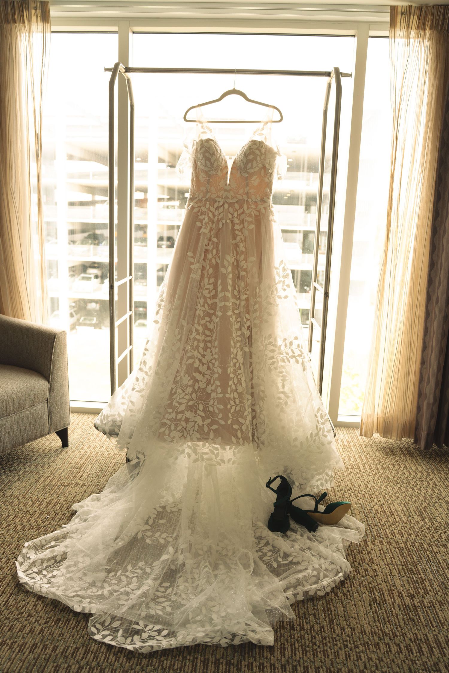 A beautiful white lace wedding dress with a long train displayed on a carpeted floor near large windows.