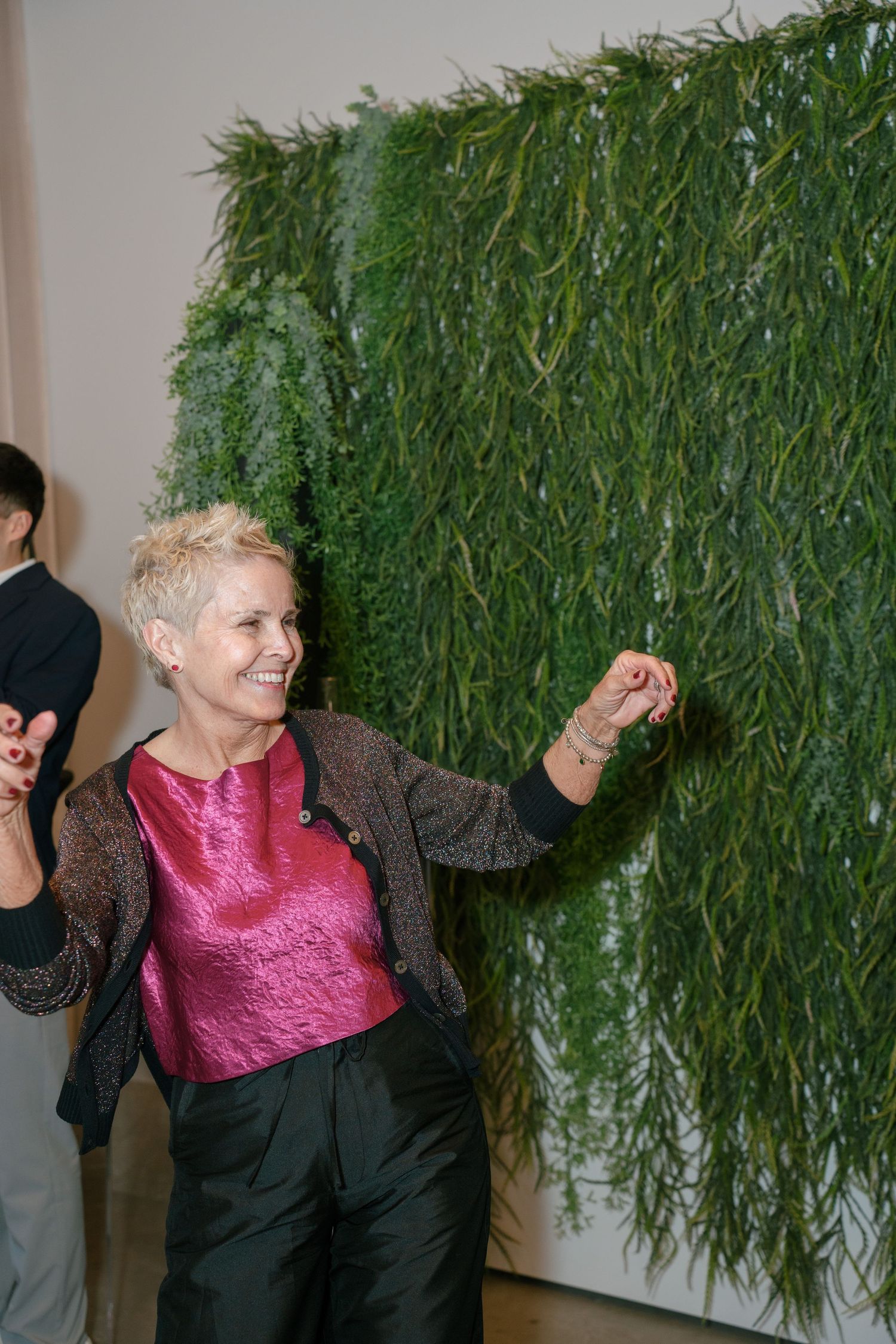 Woman in pink metallic top and cardigan dancing joyfully near green living plant wall at event.