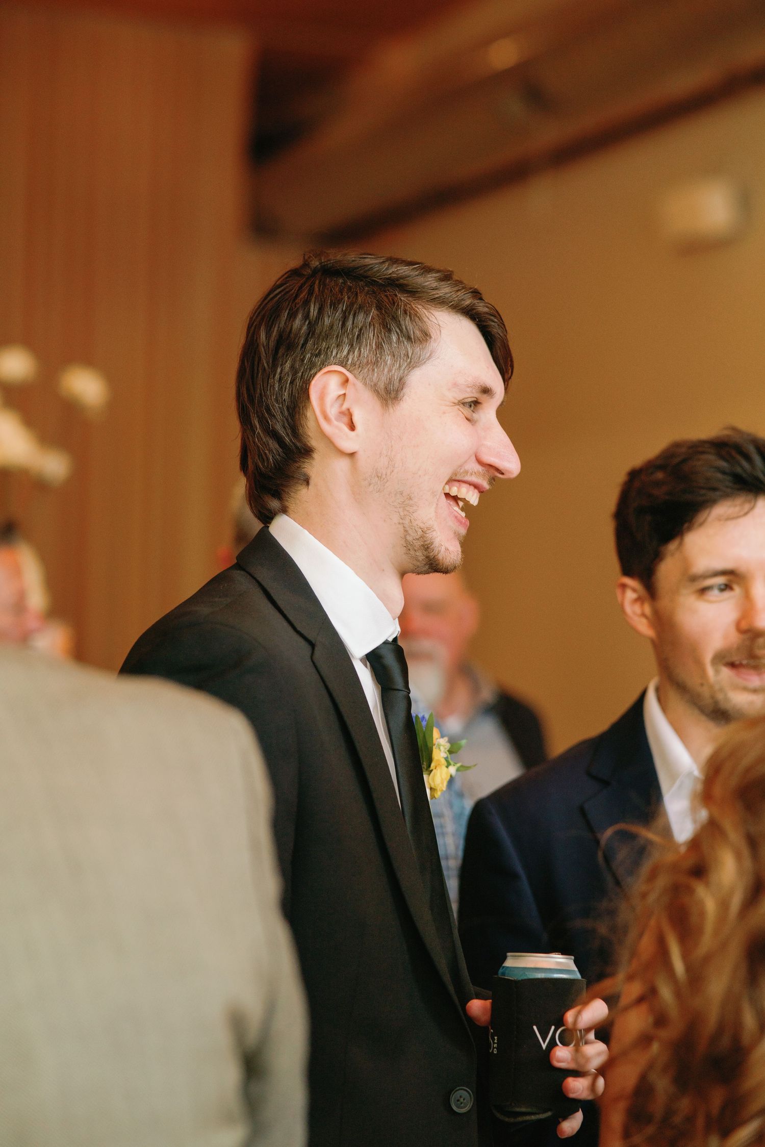 Smiling groom in dark suit with yellow boutonniere holding a coffee cup at a wedding reception.