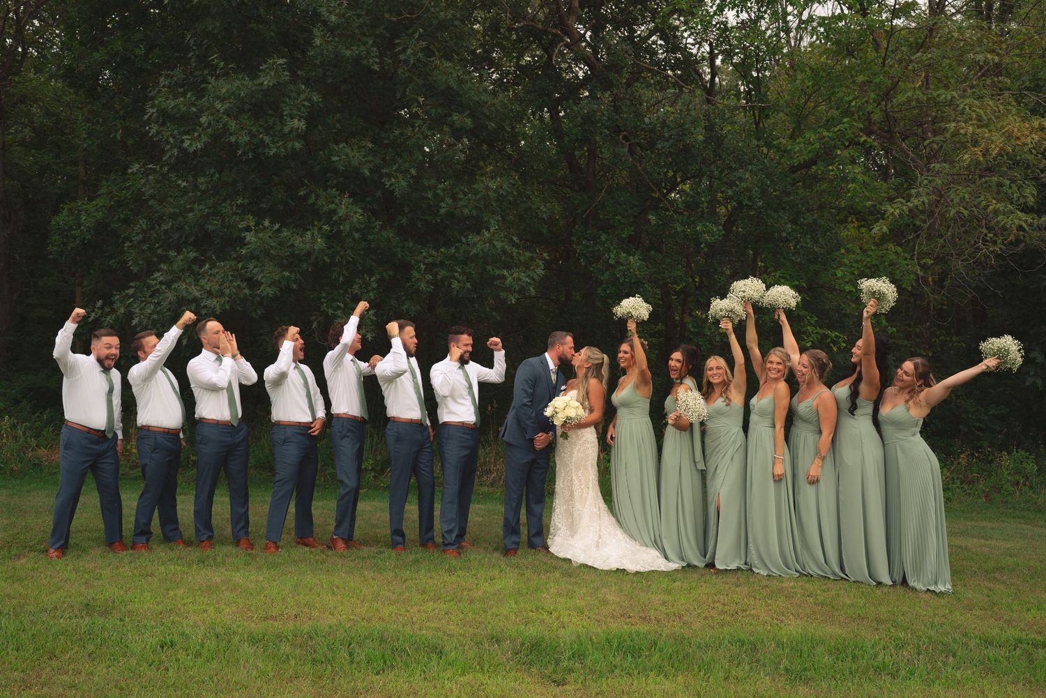 Wedding party poses in a line outdoors with bridesmaids in sage dresses and groomsmen in navy suits.