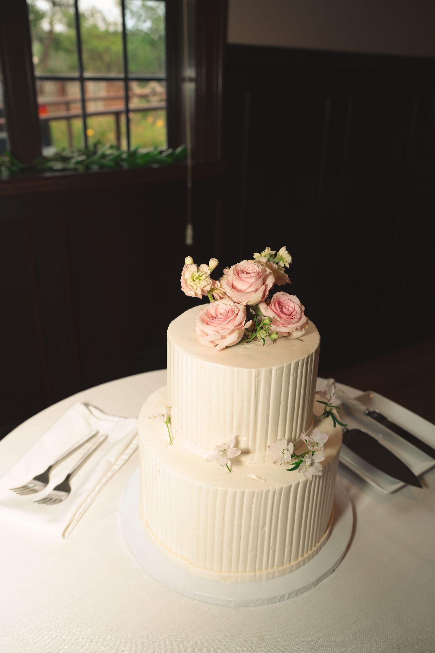 Two-tiered white wedding cake decorated with pink roses on elegant table setting.