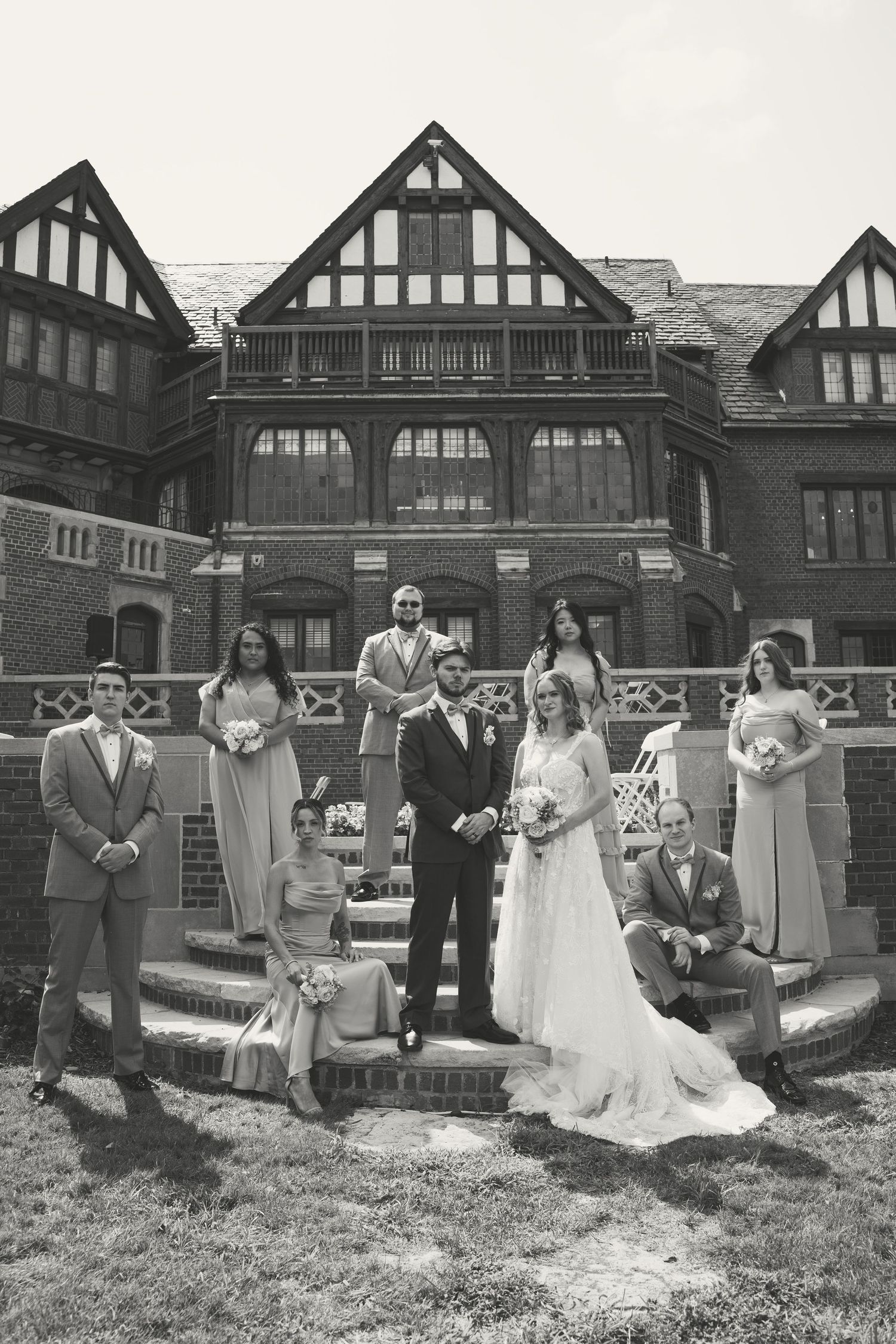Black and white wedding party photo posed on steps of elegant Tudor-style mansion with bride, groom, and attendants.