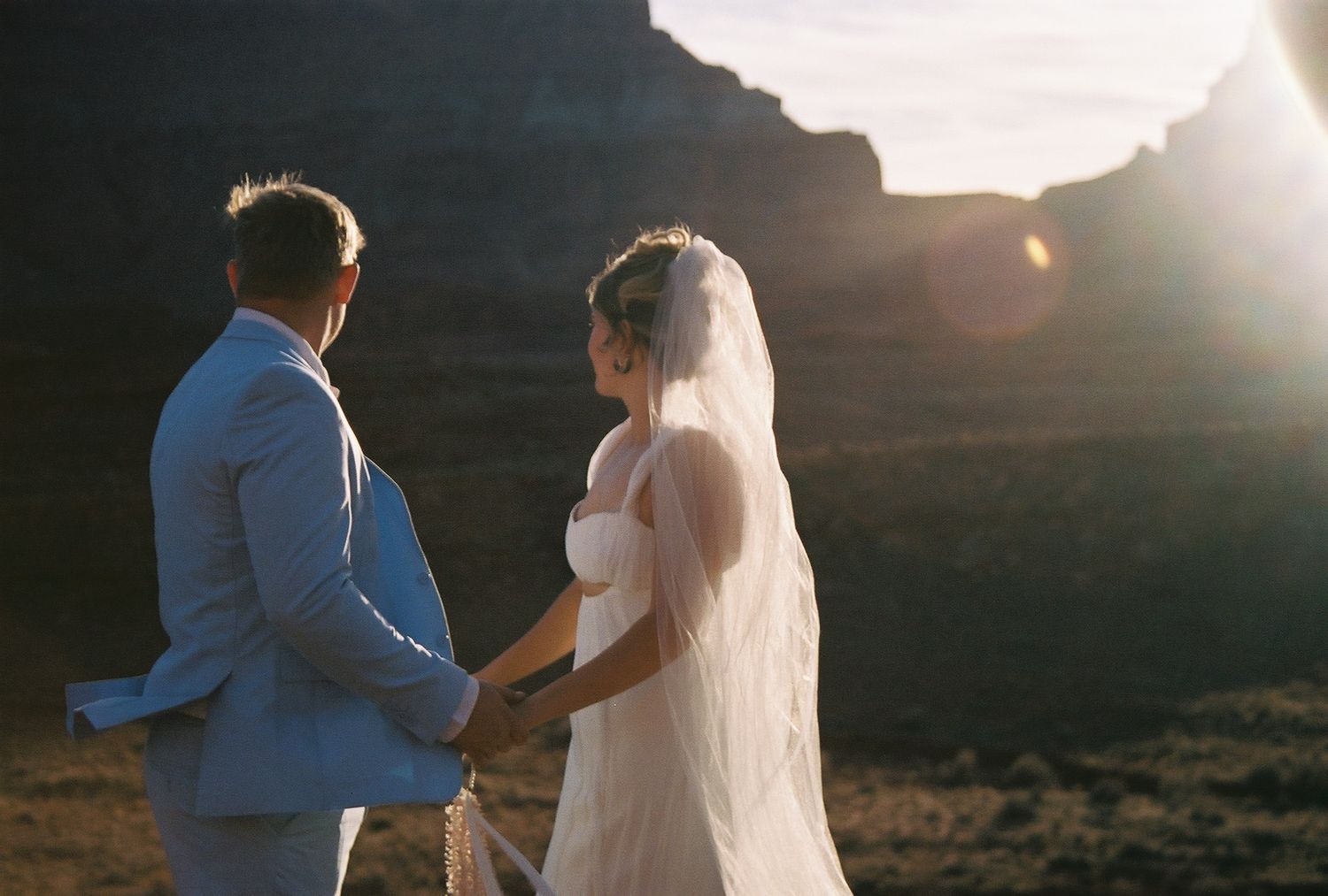 Bride and groom holding hands at sunset in a desert canyon, bride wearing white gown and veil.