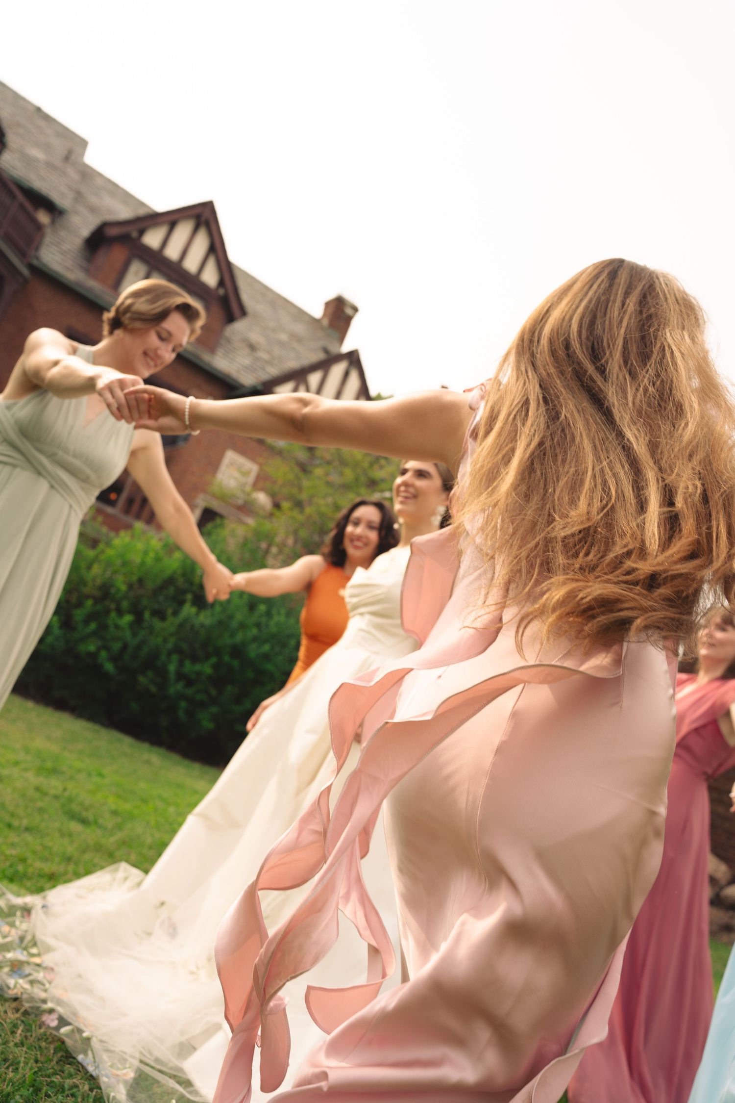 Wedding party members twirl and dance together on lawn showing flowing dresses in motion.