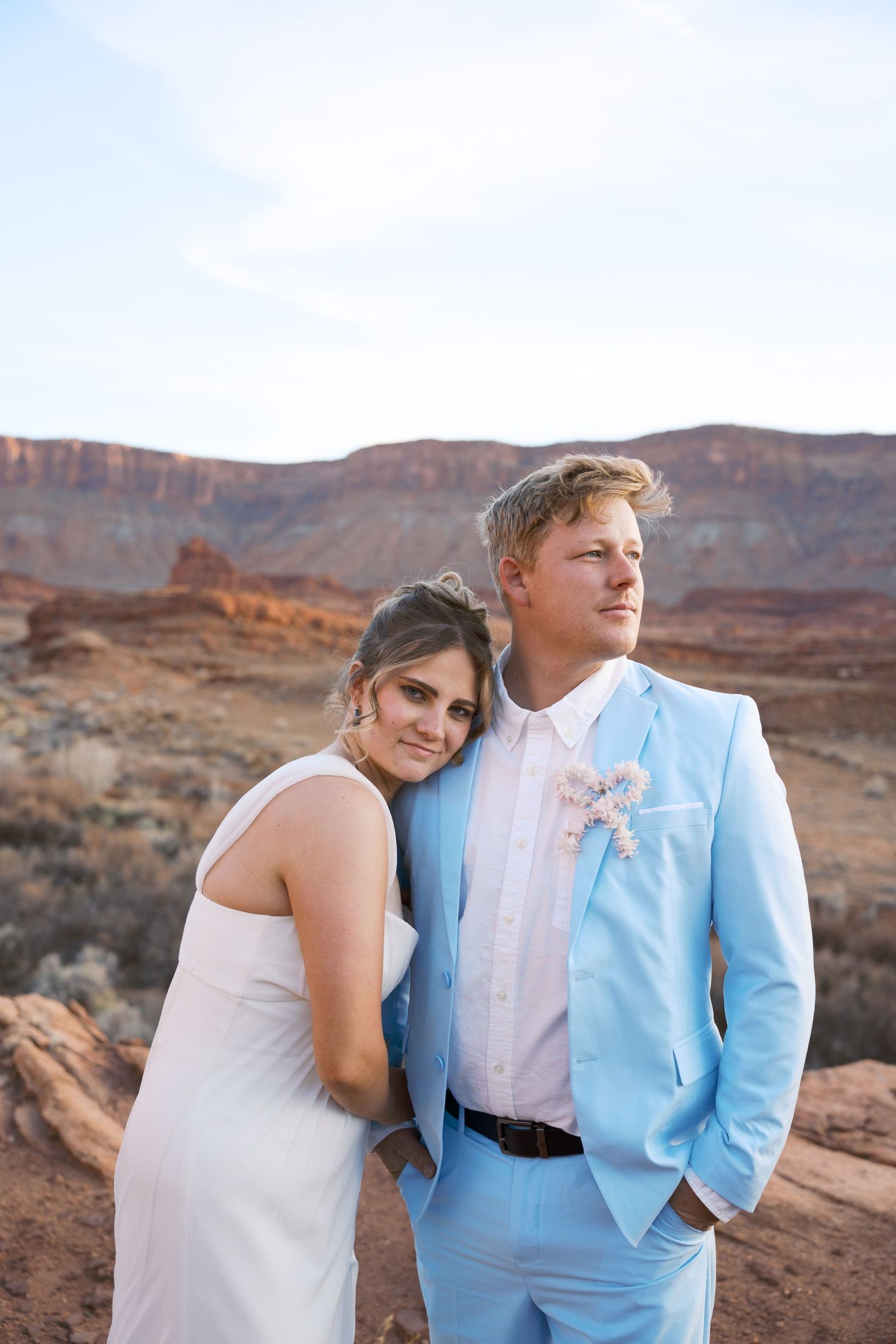 Bride and groom pose in desert landscape, she in white dress, he in light blue suit with boutonniere, red rock formations behind.