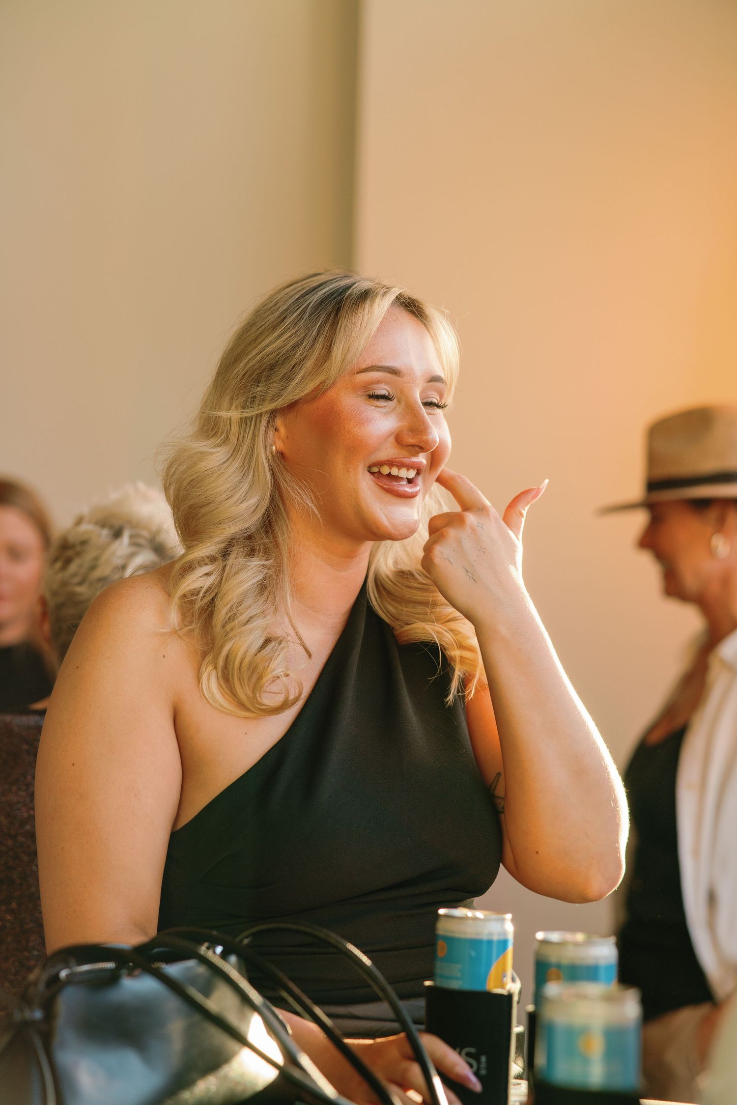 Smiling blonde woman in black dress making a phone gesture at a wedding reception.