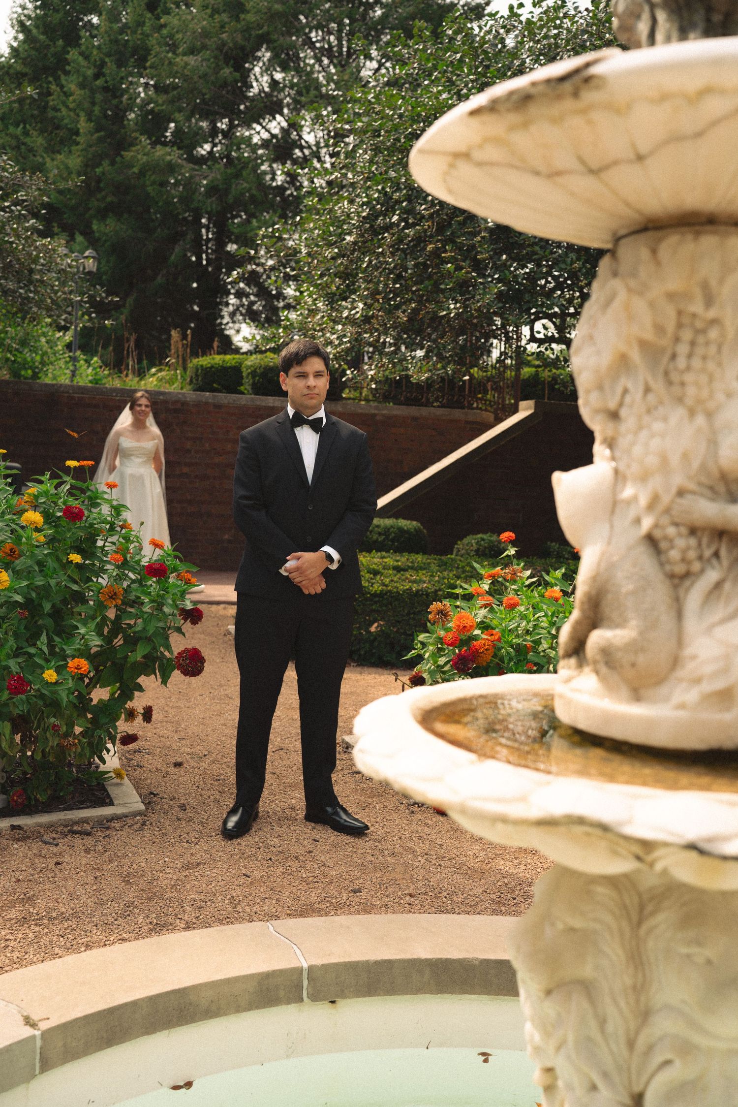 A couple poses in formal attire near an ornate fountain in a garden with orange flowers.