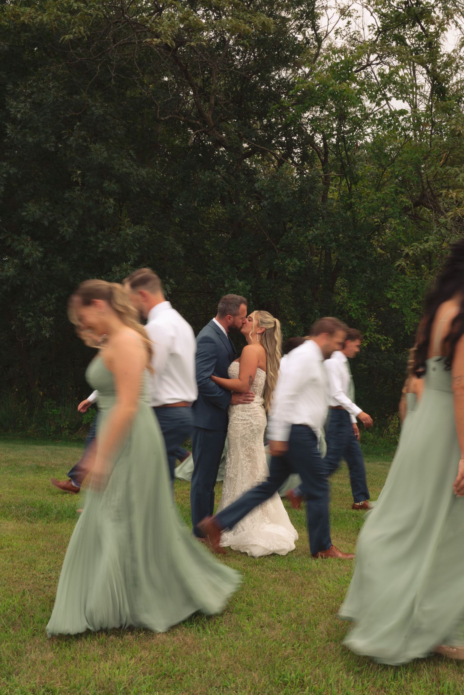 Wedding party members in sage green dresses and navy suits perform a fun falling domino effect pose on grass.