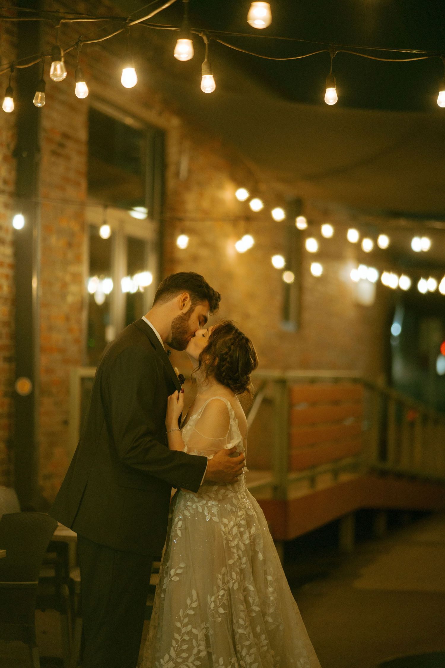 Wedding couple embracing under string lights in an industrial brick venue.