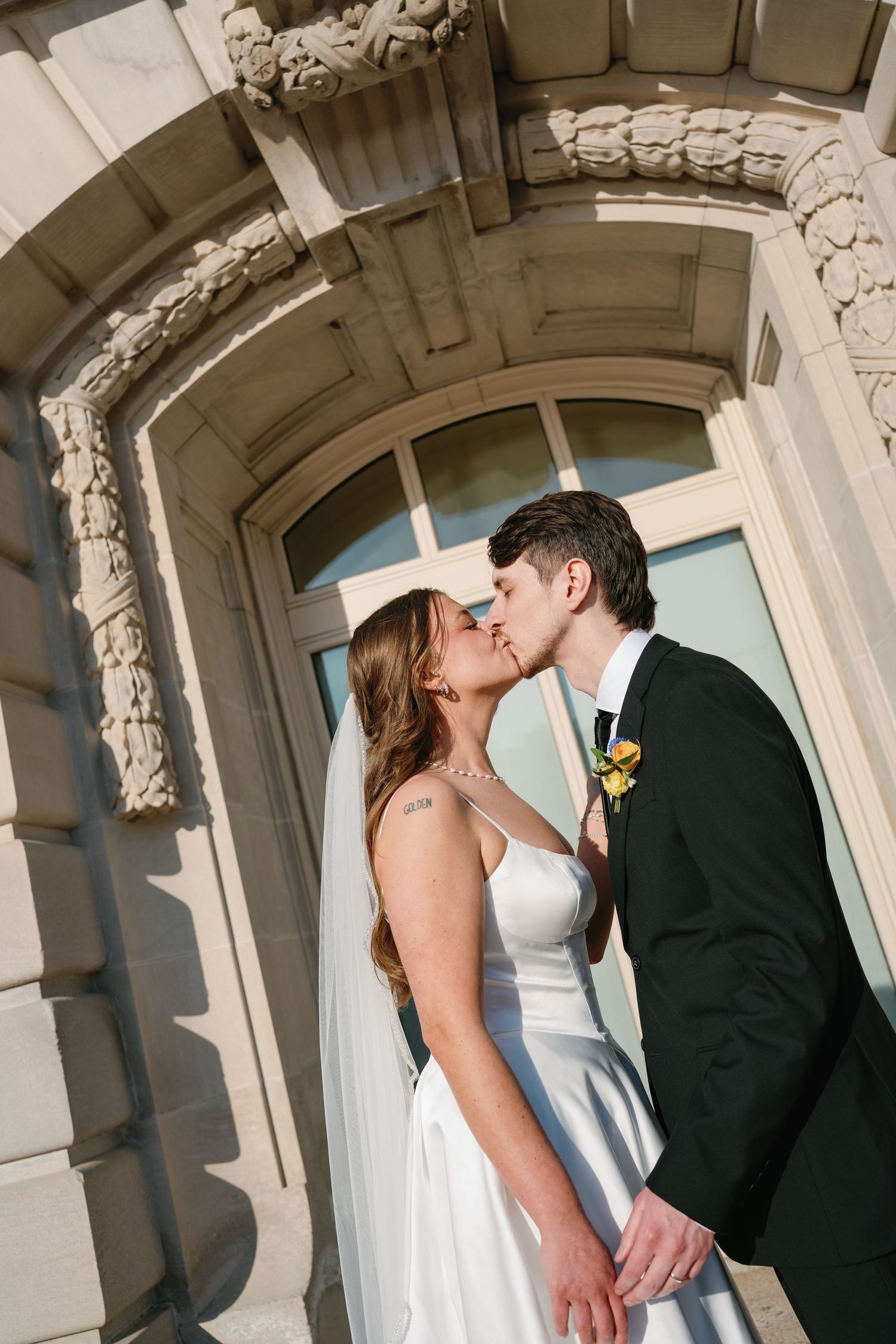 Bride and groom share romantic kiss under ornate stone archway in wedding attire.