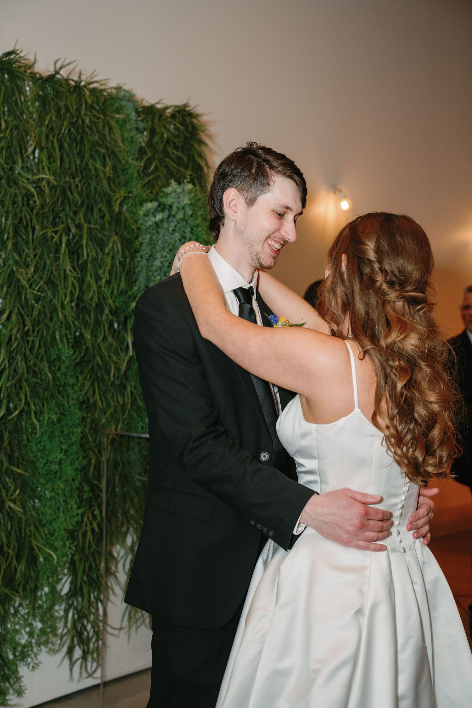 Bride and groom share their first dance, smiling, in front of a lush green wall backdrop at their wedding reception.