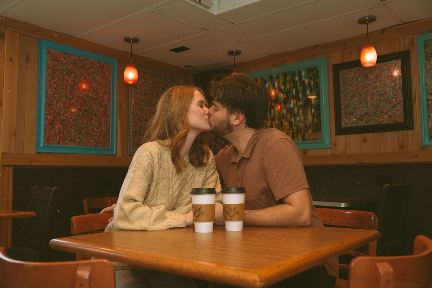 Couple sharing a kiss at a wooden cafe table with coffee cups, surrounded by colorful artwork on teal walls.