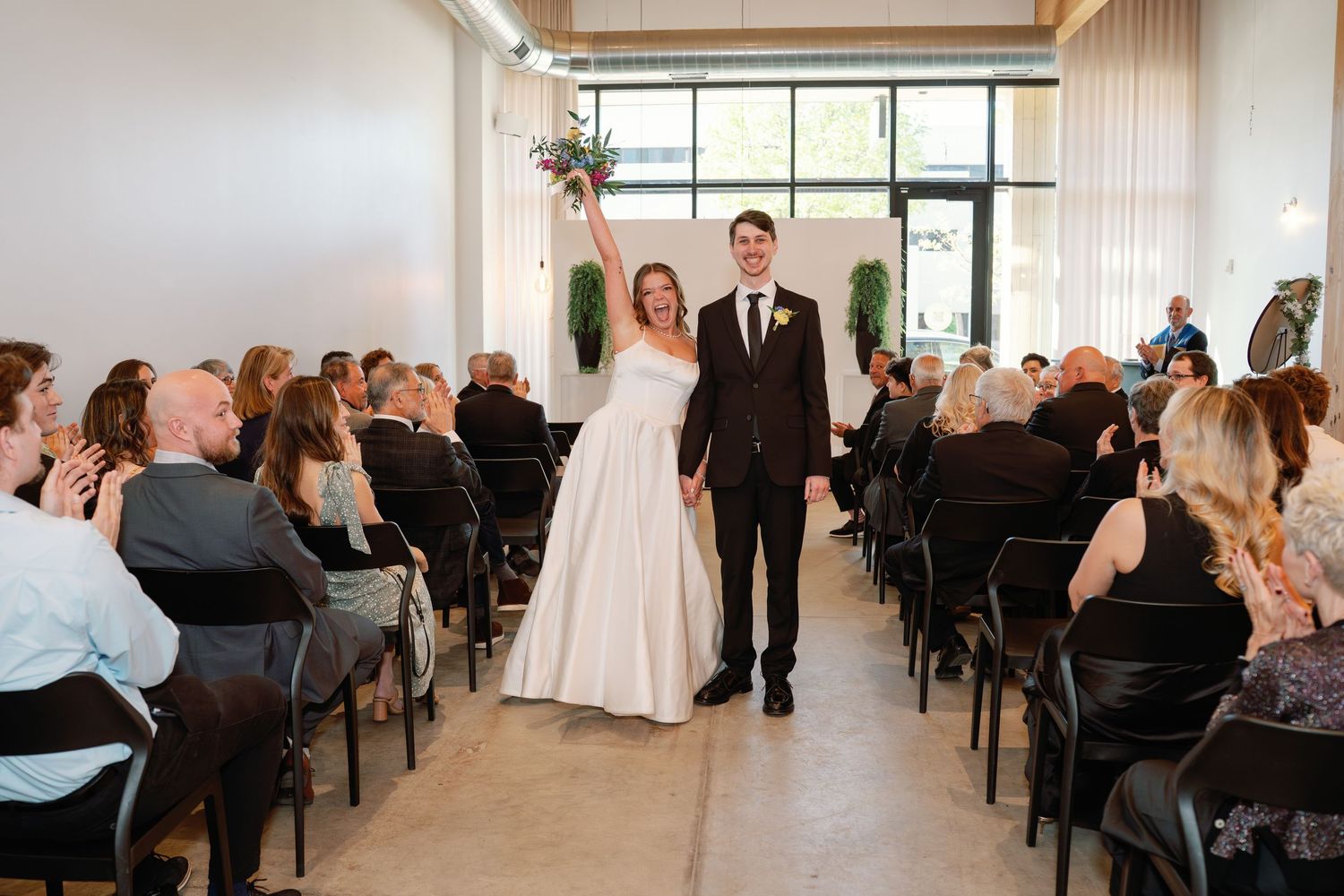 Bride and groom walk down the aisle together, smiling at guests seated in a modern wedding venue.