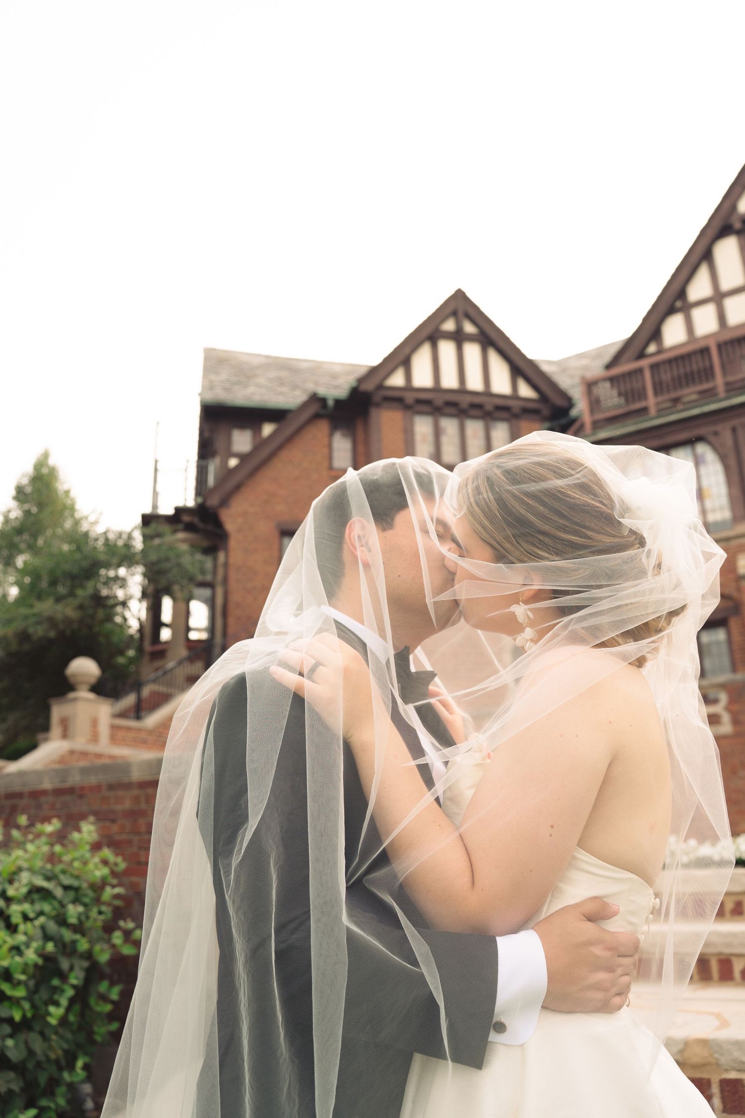 Romantic wedding couple sharing a kiss under a flowing veil in front of a Tudor-style mansion.