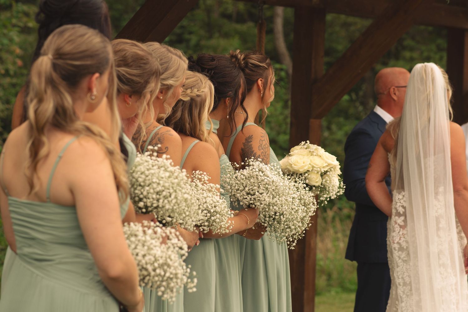 Wedding ceremony participants gather under a wooden pavilion surrounded by greenery.