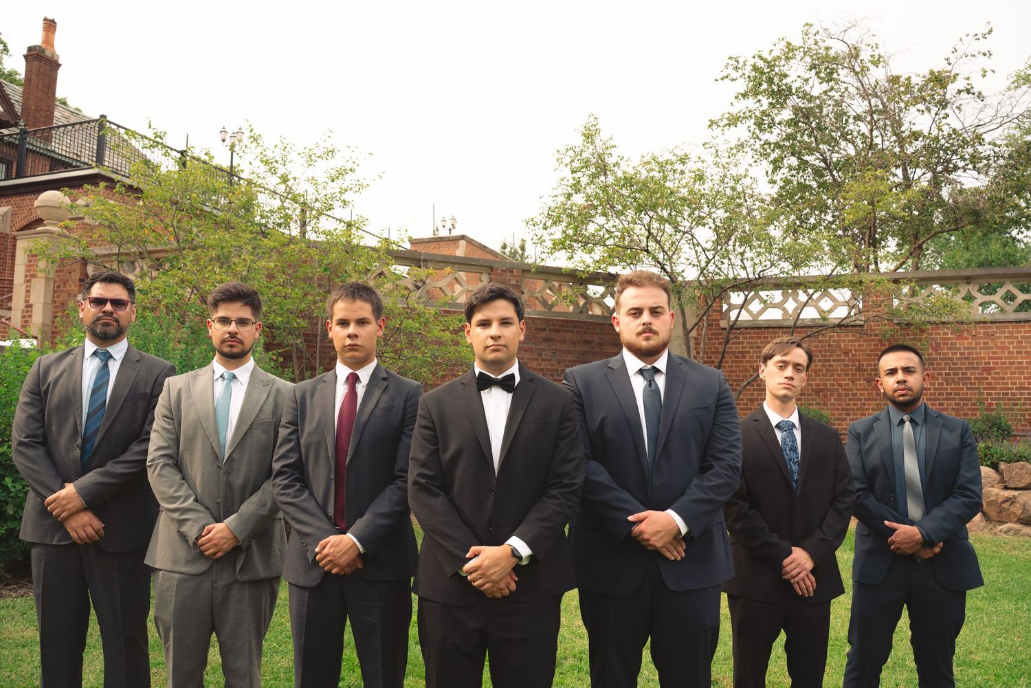 Groomsmen in dark suits and ties stand in formation on grass outside a brick building.