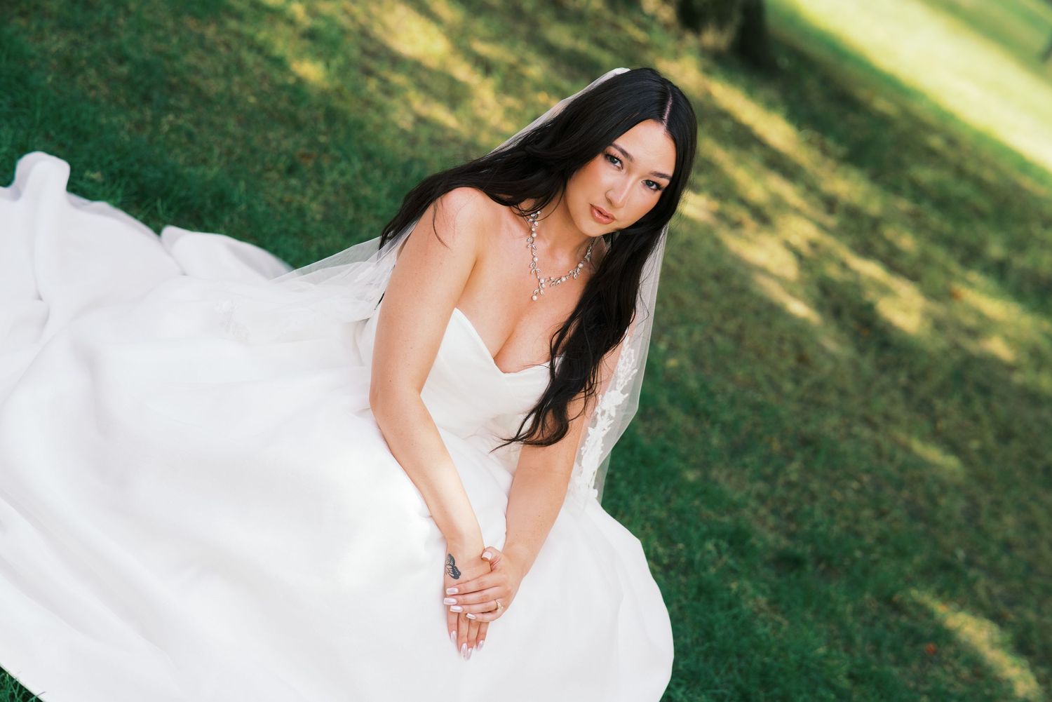 Bride in white strapless gown sitting on green grass, long dark hair, elegant necklace, outdoor wedding portrait.